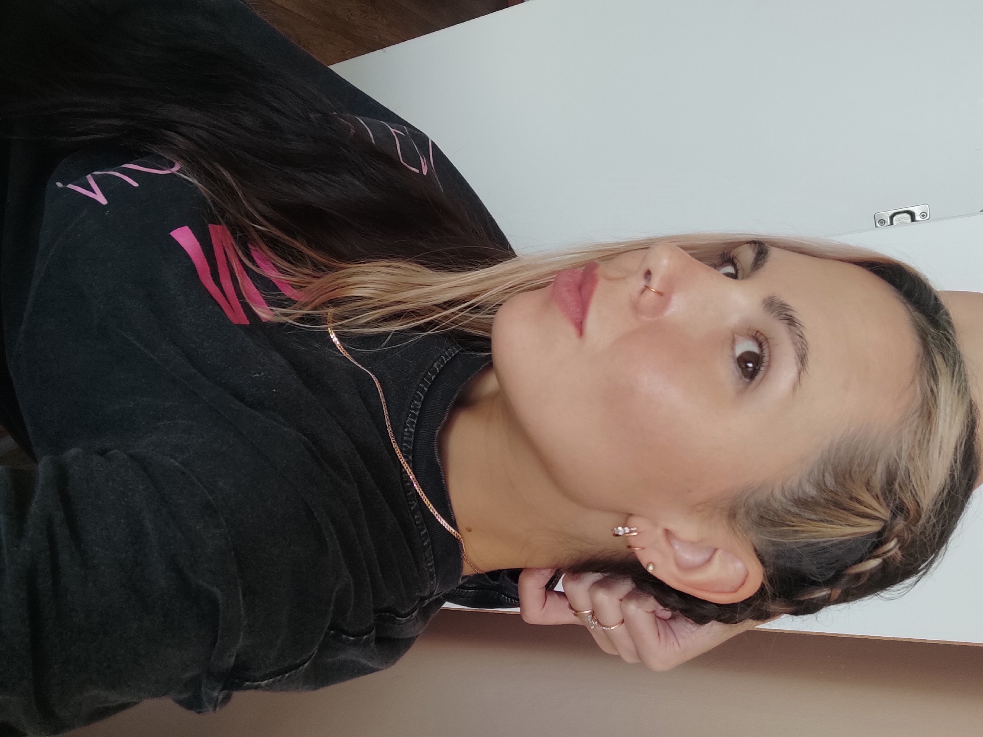 A woman is lying on her side, facing the camera. She has dark hair with blonde highlights, styled in a braid along her hairline. She is wearing a dark-colored sweatshirt with pink lettering. She has a nose ring and several earrings. A thin gold necklace is visible around her neck. Her hand is near her ear with several rings. The background includes a white wall and a brown surface. There is no indication that the image was taken in Chaikovsky, Russia.