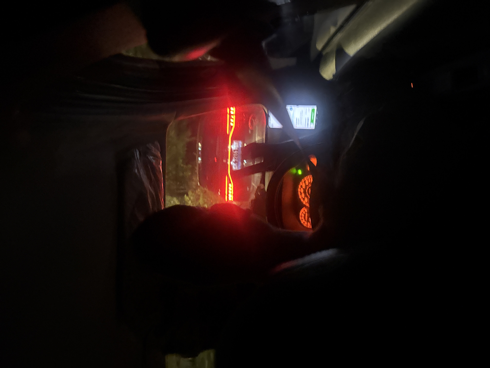 The media depicts the dark interior of a vehicle at night, likely captured from a passenger's perspective, looking towards the front and out the windshield. The scene is dominated by artificial light sources against the darkness.
In the foreground, a person's arm and shoulder are partially visible on the left, and another person's head or shoulder is dimly outlined on the far right, suggesting at least two occupants. A prominent vertical strip of bright red LED lights runs through the center of the image, possibly an interior accent light or a reflection of external vehicle lights. Through the windshield or side window, faint green foliage can be seen, illuminated by the red glow, hinting at an outdoor, possibly urban or suburban, setting.
A smartphone is mounted near the windshield on the right side, its screen brightly illuminated with a user interface that features significant green elements. This appears to be a navigation or ride-sharing application. While most text is illegible due to distance and brightness, vague shapes resembling numbers like "12.05" or "12.25" and a letter "A" can be discerned on the screen, along with other icons and what looks like a map display. To the right of the phone, a vehicle dashboard or instrument panel glows with orange and red lights, revealing circular gauges. A plastic bag is visible on the left side of the vehicle's interior.
The overall impression is of nighttime travel in a vehicle, possibly a taxi or ride-share, within the Kuta Selatan, Indonesia area, given the contextual information. No specific weather conditions are discernible, and no explicit emotions are evident.