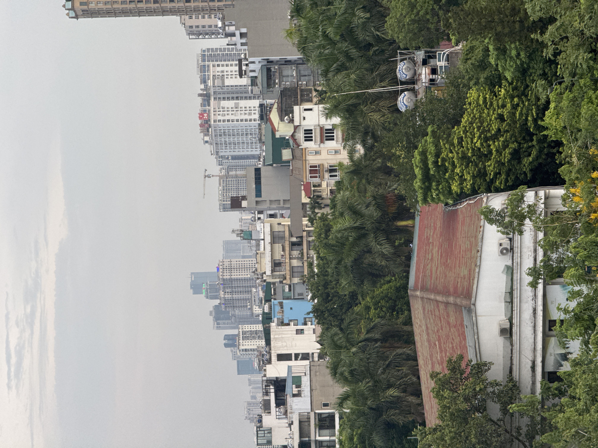 This media file captures a cityscape in Hà Nội, Vietnam, viewed from an elevated perspective. The scene is dominated by a dense urban environment with numerous buildings of varying heights and architectural styles, interspersed with lush green trees. In the foreground, a building with a rusty, corrugated metal roof is partially visible, adjacent to a white structure with visible air conditioning units.
Further into the cityscape, a dense collection of high-rise buildings stretches across the horizon, some appearing modern and glass-fronted, while others are older, multi-story structures. Construction cranes are visible among the buildings, indicating ongoing development. The sky is overcast and hazy, suggesting a cloudy or potentially polluted day, and it is difficult to determine the exact time of day, though it appears to be daytime.
Towards the right side of the frame, partially obscured by trees, are elements of what appears to be a small amusement park or recreational area, including what look like two large, enclosed circular structures that could be Ferris wheel cabins. Text is visible on these circular structures; it appears to be a logo or branding. One visible word on the blue circular elements is "SONG".
The overall impression is one of a bustling, densely populated city with a mix of the natural and built environments. There are no people prominently featured, and the scene is static, without any discernible activity or event taking place. The weather appears to be humid and overcast.