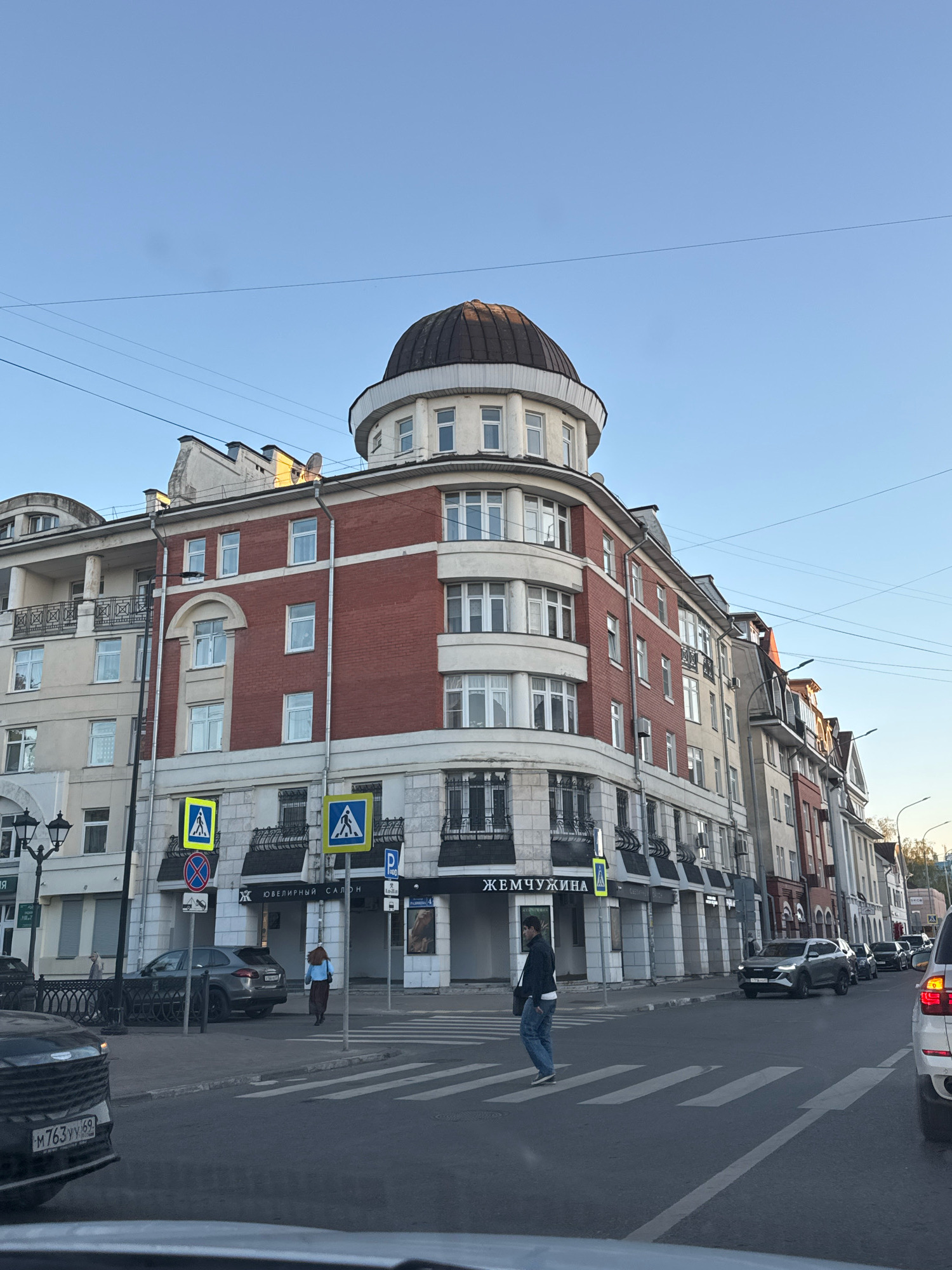 The image depicts a street scene in Tver, Russia, under a clear blue sky. A multi-story building with a distinctive domed structure at the top is prominent on the right side of the frame. The building features red brick on its upper levels and white stone or stucco on the lower levels. Signs are visible on the building, including one that reads "ЖЕМЧУЖИНА" (Zhemchuzhina).
Several pedestrians are present in the scene. One individual is crossing a crosswalk in the foreground. Another person is walking on the sidewalk near the building. Cars are on the street, including a dark SUV in the middle ground and a white SUV in the foreground, partially obscuring the view. Traffic signs, including pedestrian crossing signs, are visible at the intersection. The environment appears to be an urban setting with streetlights and power lines also visible. The overall lighting suggests it is late afternoon or early evening.