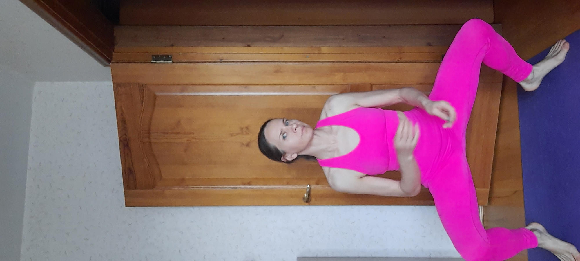 An adult female, likely in Moscow, Russia, is shown indoors performing a yoga or stretching pose. She is wearing a striking, solid hot pink athletic ensemble, including a sports bra or crop top and full-length leggings. Her dark hair is pulled back, and she looks directly at the camera with a neutral and focused expression.
She is positioned against a warm, natural-toned wooden surface, possibly a wardrobe or a large wooden door, which forms the majority of the background. Her body is arranged horizontally across the frame, with her legs spread wide apart. Her bare feet rest on a deep purple exercise mat that extends to the right side of the image. Her arms are bent, with her hands positioned near her hips or abdomen. To the left of the wooden surface, a light-colored wall with a subtle, textured pattern is visible. The setting suggests a home environment. No text is visible in the image.