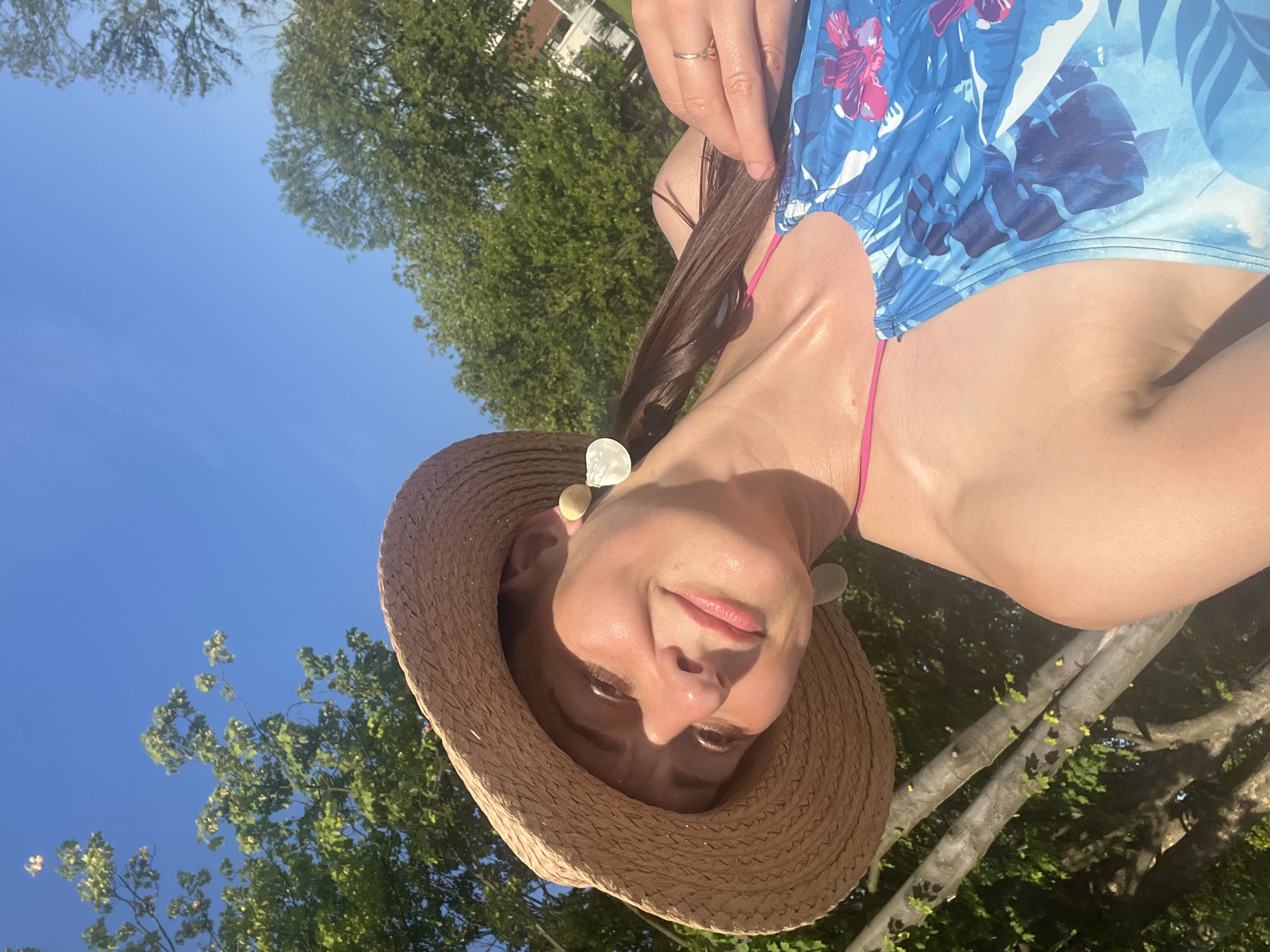 In a close-up, low-angle selfie taken outdoors, a woman with a straw hat tilted downwards smiles at the camera. Her hair, pulled back, is visible beneath the hat. She wears a blue swimsuit with a tropical floral pattern, and a pink strap is visible. She also wears a ring on her right hand, and large, light-colored earrings. The background consists of a bright blue sky and lush green trees. Sunlight casts shadows on her face, highlighting her features. The image appears to be taken in a natural, possibly backyard, setting.