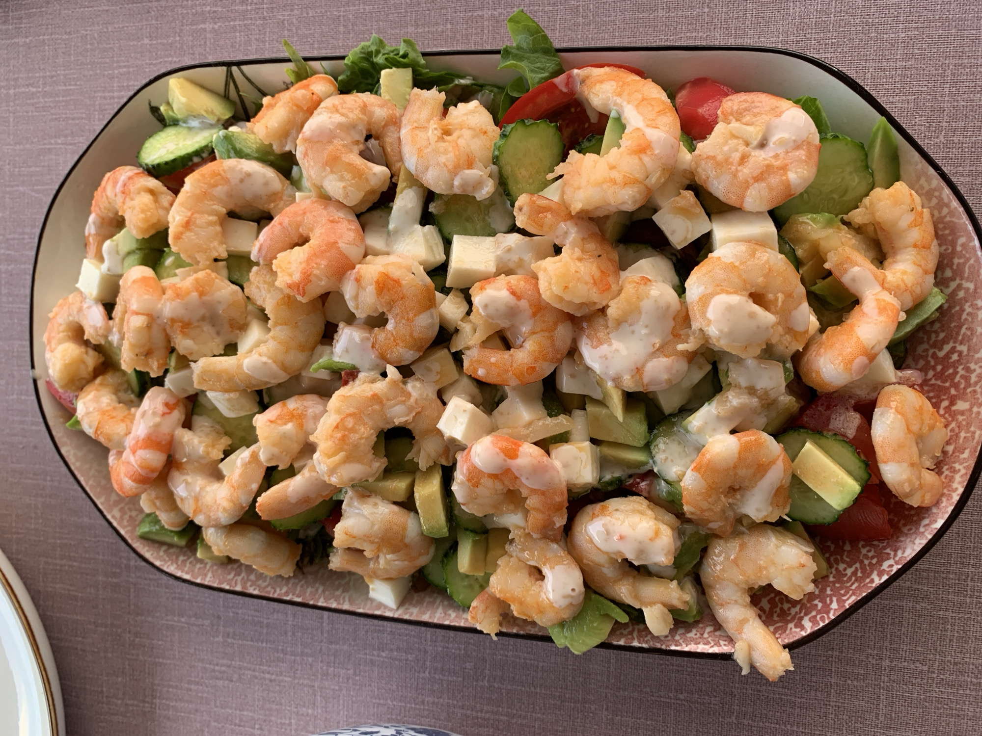 An overhead shot shows a large, oval plate filled with a shrimp salad. The salad is comprised of several ingredients, including cooked shrimp, diced avocado, cubes of white cheese, sliced cucumber, and red cherry tomatoes. The shrimp are plump and pink with a creamy dressing drizzled over them. The plate itself has a pink and white patterned rim. The salad rests on a textured, pinkish-grey tablecloth. The background shows a sliver of a white plate with a gold rim. The image is well-lit, focusing on the food. There is no indication of the city of Суроватиха, Russia, or any people present in the image.