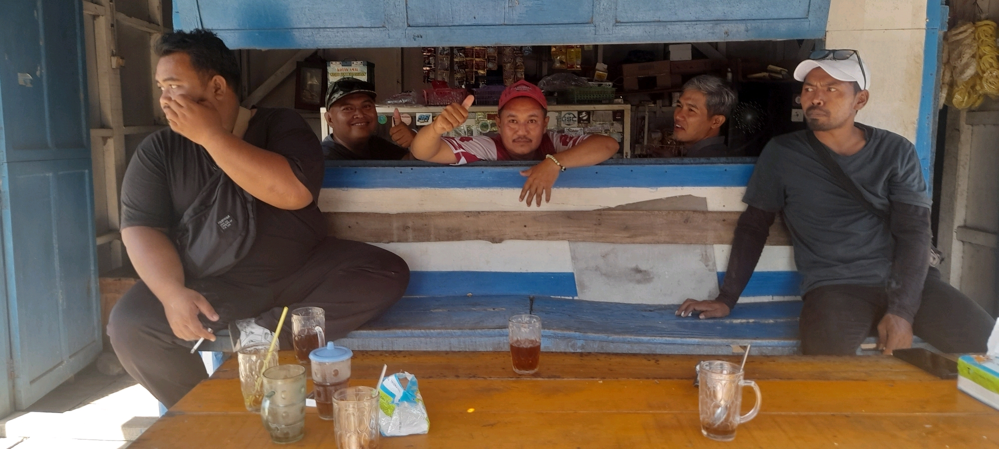 In Sokawati, Indonesia, five men are gathered around a wooden table outside a small open-air shop. The man on the far left, who is large, wears a black t-shirt and dark pants, with a black fanny pack across his chest. He is looking to his left with his hand near his face. To his right, inside the shop, two men are visible behind a counter. The man in the middle of the shop wears a camouflage cap and a dark shirt, smiling and giving a thumbs-up. Next to him, a man in a red cap and a red and white striped shirt leans forward, also giving a thumbs-up and looking towards the camera. Further to the right, a man with graying hair and a dark shirt looks to his left. On the far right, a man wearing a white baseball cap backwards and sunglasses on top, a dark t-shirt, and dark pants, has a black bag slung across his chest and is looking to his right. The table in front of them holds several glasses of a dark beverage, a plastic bag, and a cigarette. The shop behind them appears to sell snacks and drinks.