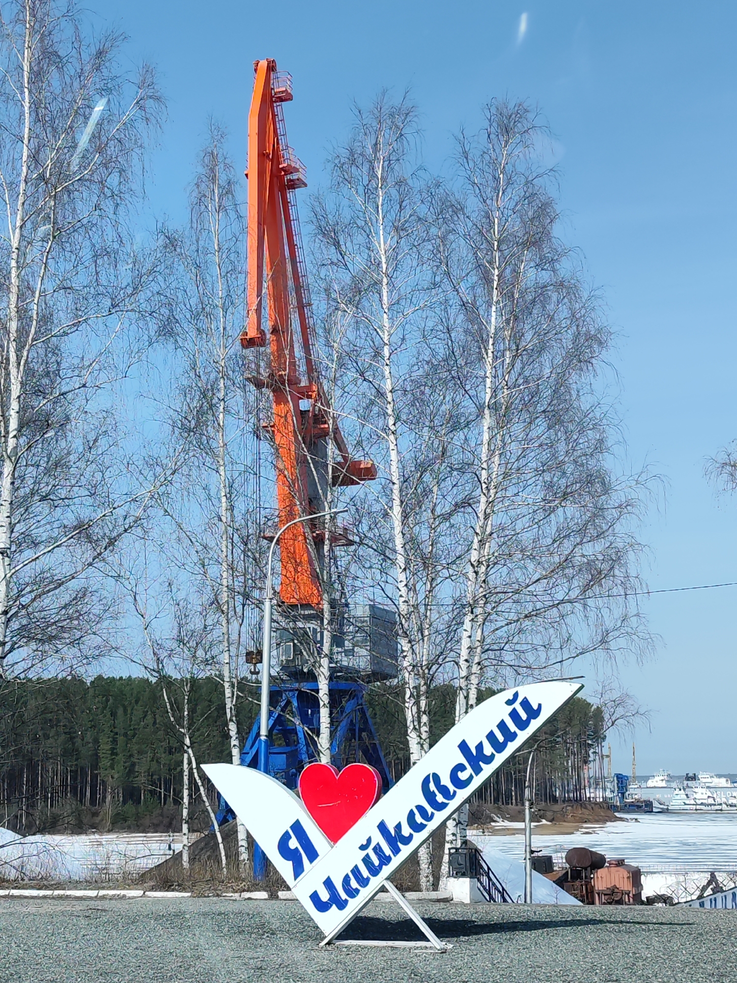 A daytime shot shows a sign at the entrance to the city of Chaikovsky, Russia, featuring the words "I love Chaikovsky" written in Russian, next to an orange gantry crane behind leafless birch trees in the background. The sign is white with blue lettering and a red heart. Behind the trees is a forest and a partially frozen body of water. The sky is clear and blue. The crane's base is blue and gray. The ground in front of the sign is covered with small stones. Parts of boats and other structures are seen in the background.