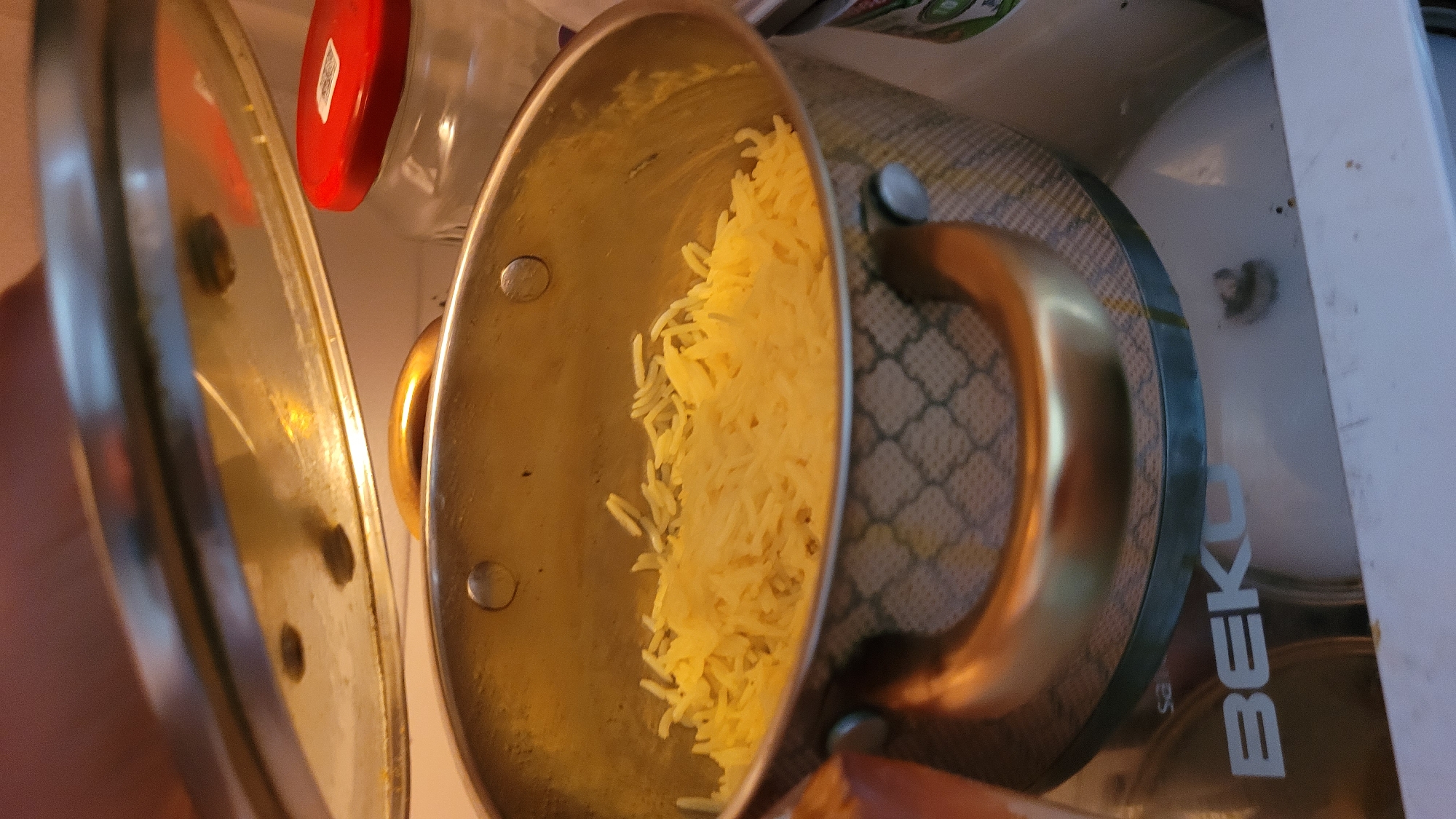 A top-down, close-up view shows a stainless steel pot filled with cooked rice, likely basmati, sitting inside a refrigerator in Unknown, Unknown. A stainless steel lid, slightly ajar, rests to the left of the pot. Adjacent to the pot are various items including plastic containers, one with a red lid, and what appears to be a canister with the word "BEKO" visible on its side. The interior of the refrigerator is white. The rice appears to be yellowed, possibly from butter or saffron. The pot has gold-colored handles.