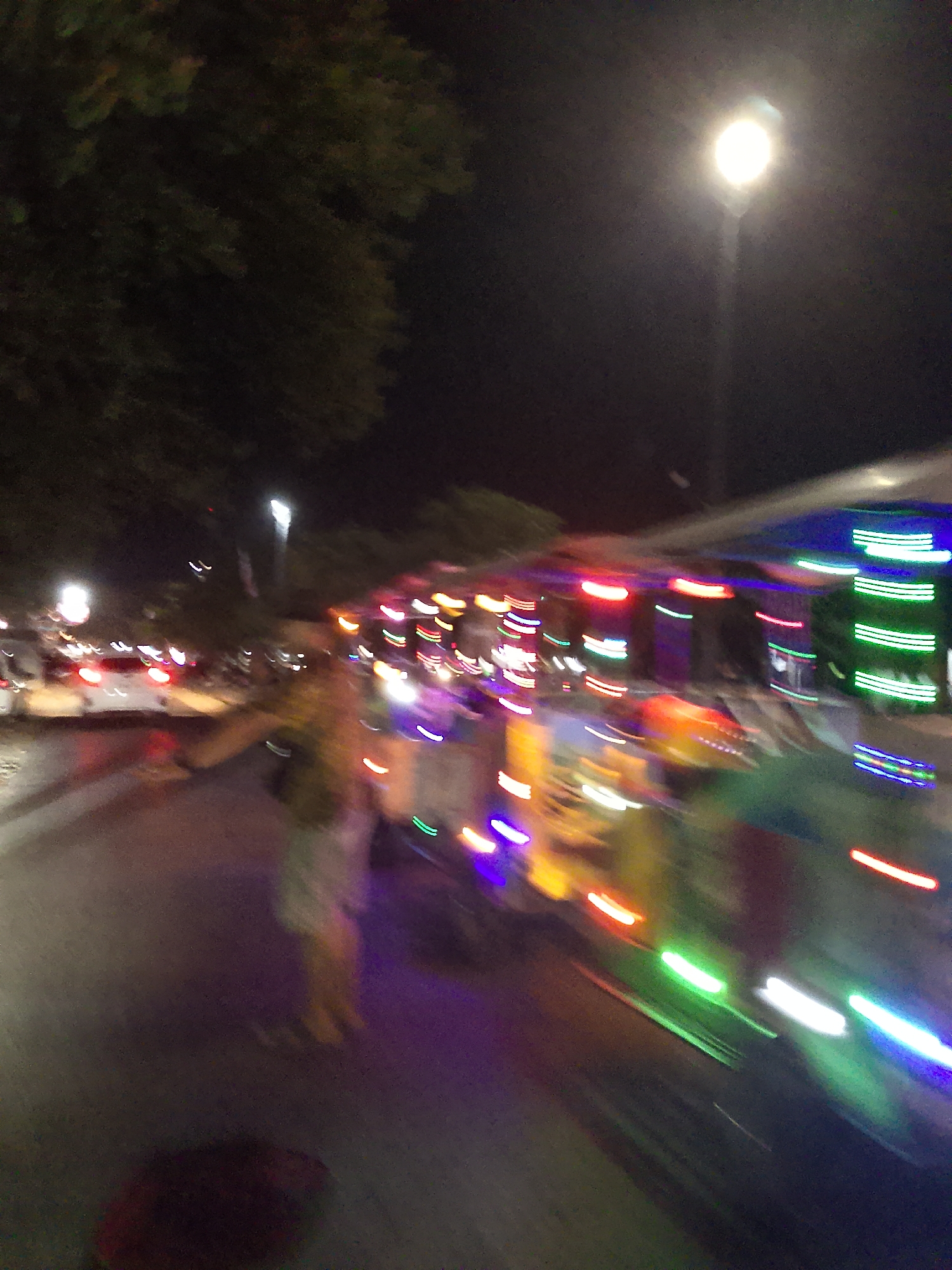 The image captures a street scene at night in Balun, Indonesia, characterized by motion blur due to movement. A vehicle, possibly a food cart or a small truck, is visible with an array of colorful lights, including green, red, and blue, decorating its exterior. The lights are streaked across the image, indicating rapid motion. In the background, trees are dimly lit by streetlights, one of which is prominently visible in the upper right quadrant of the image, emitting a bright white glow. The street itself is dark, with the primary illumination coming from the vehicle's lights and the street lamp. The overall impression is a dynamic and lively nighttime atmosphere, possibly from a festival or a busy market area.