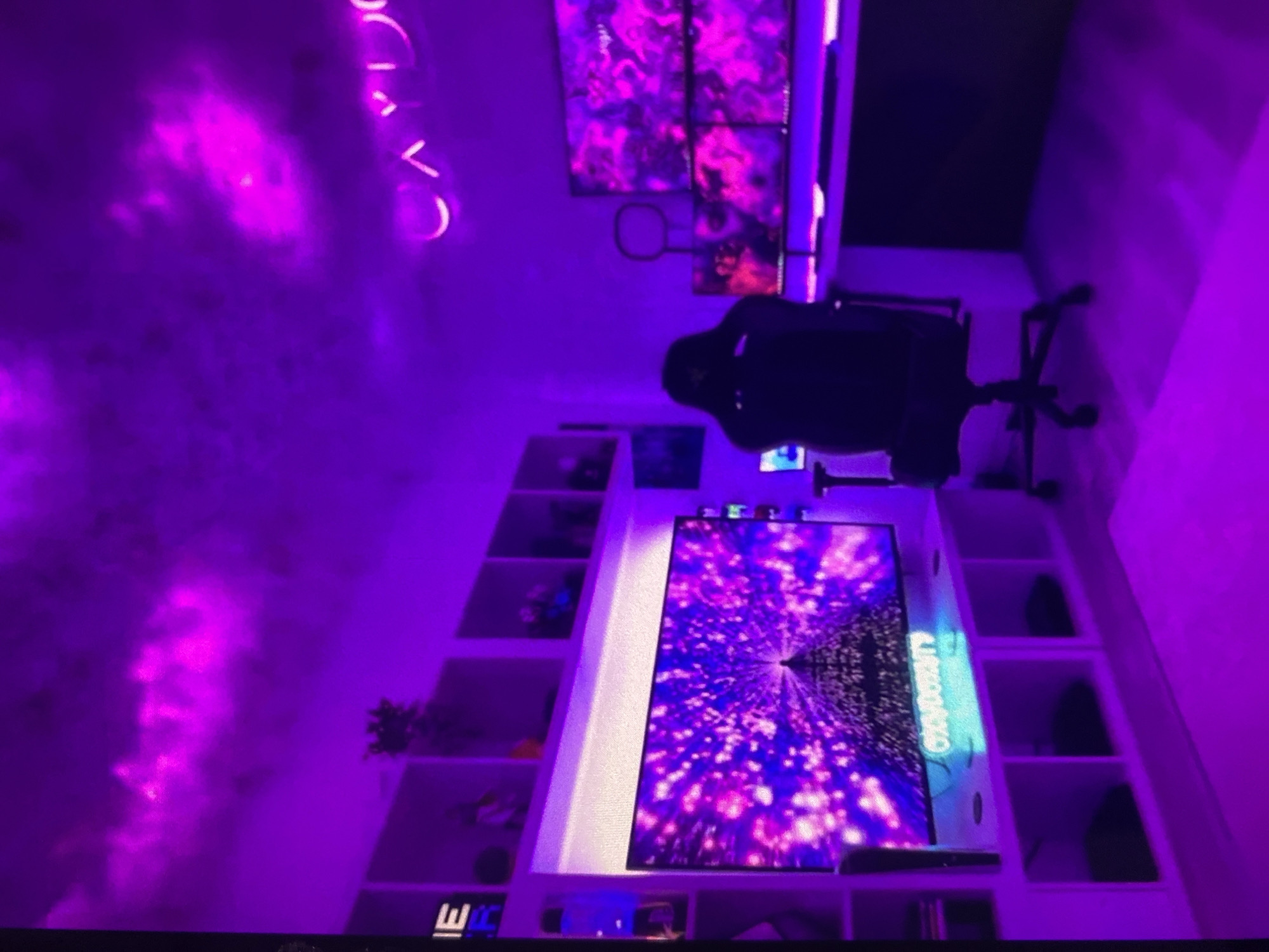 The image depicts an interior space, possibly a gaming room, characterized by purple lighting. A gaming chair is positioned in front of a setup consisting of multiple screens, some arranged in a grid-like configuration on the wall and one larger screen embedded within a shelf unit. The larger screen displays an image of a purple vortex-like tunnel. The shelves are filled with various items, including plants and decorative objects. On the wall, there's a neon sign. The location is unknown and cannot be determined from the image alone, so location information for Lhokseumawe, Indonesia is irrelevant.
