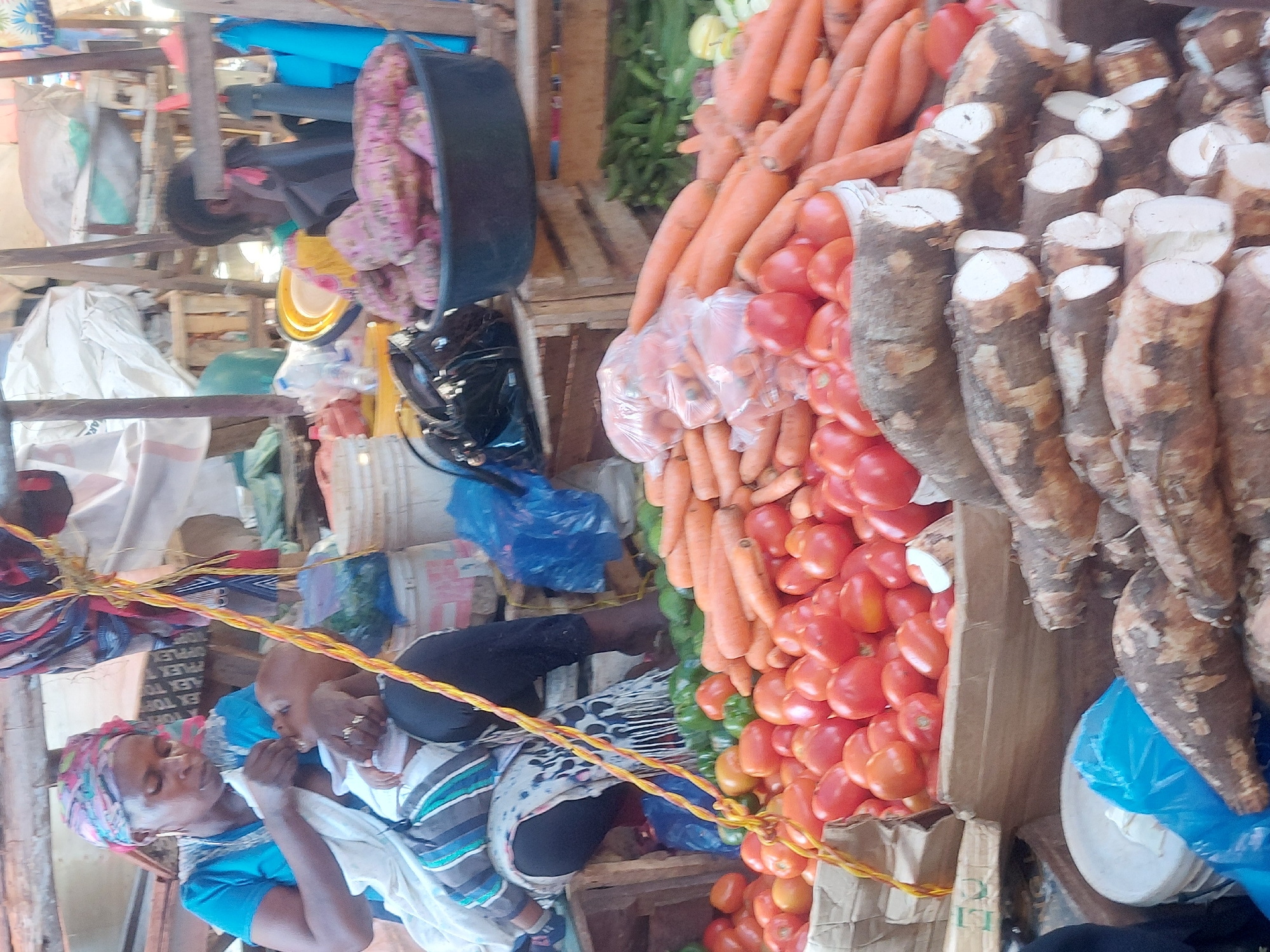 The image captures a bustling market scene in Dar es-Salaam, Tanzania. A woman wearing a blue shirt and a colorful headscarf is breastfeeding a baby. They are seated amongst piles of produce: tomatoes, carrots, cassava, and green peppers. There are also bags and bins in the background with other items and more produce. The environment is a typical market setting with wooden stalls. The image depicts a snapshot of daily life and commerce in the city.