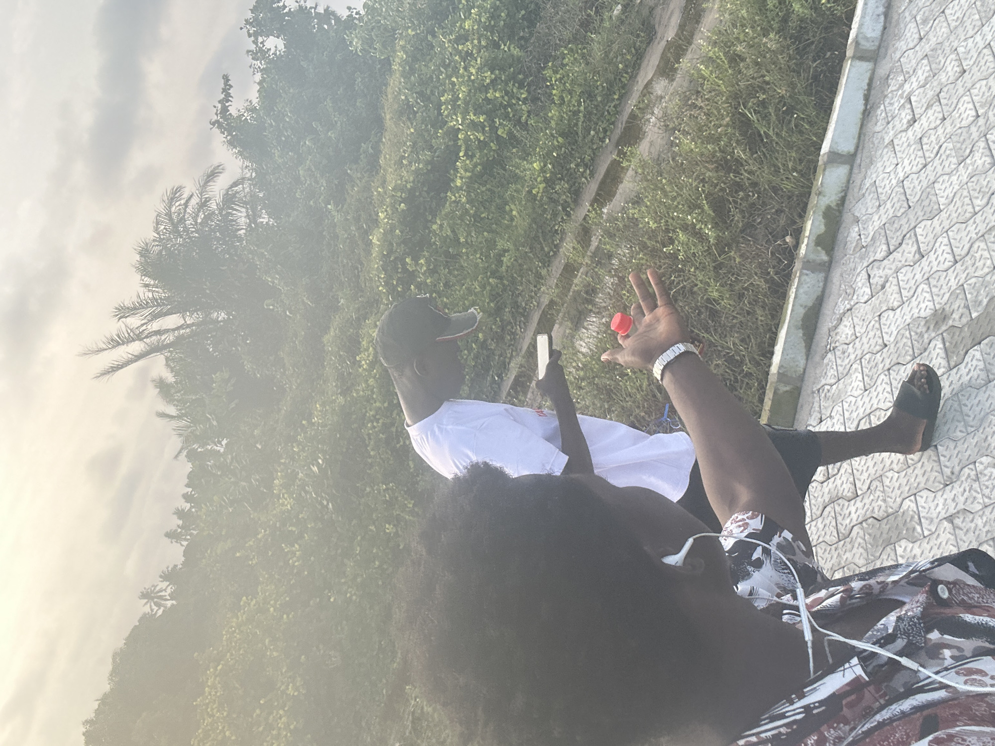 In Badagry, Nigeria, two young men are captured in a candid moment outdoors. The foreground features a person with dark, curly hair, wearing a patterned shirt and white earphones, their left hand extended with a red-capped bottle held aloft. In the mid-ground, a man in a white t-shirt and a black cap is looking down at a phone. They are situated on a paved walkway with a subtle pattern, bordered by lush green vegetation. The background reveals a dense tropical landscape with palm trees against a hazy, overcast sky, suggesting a warm, humid environment characteristic of the region. The overall scene evokes a relaxed, casual atmosphere.