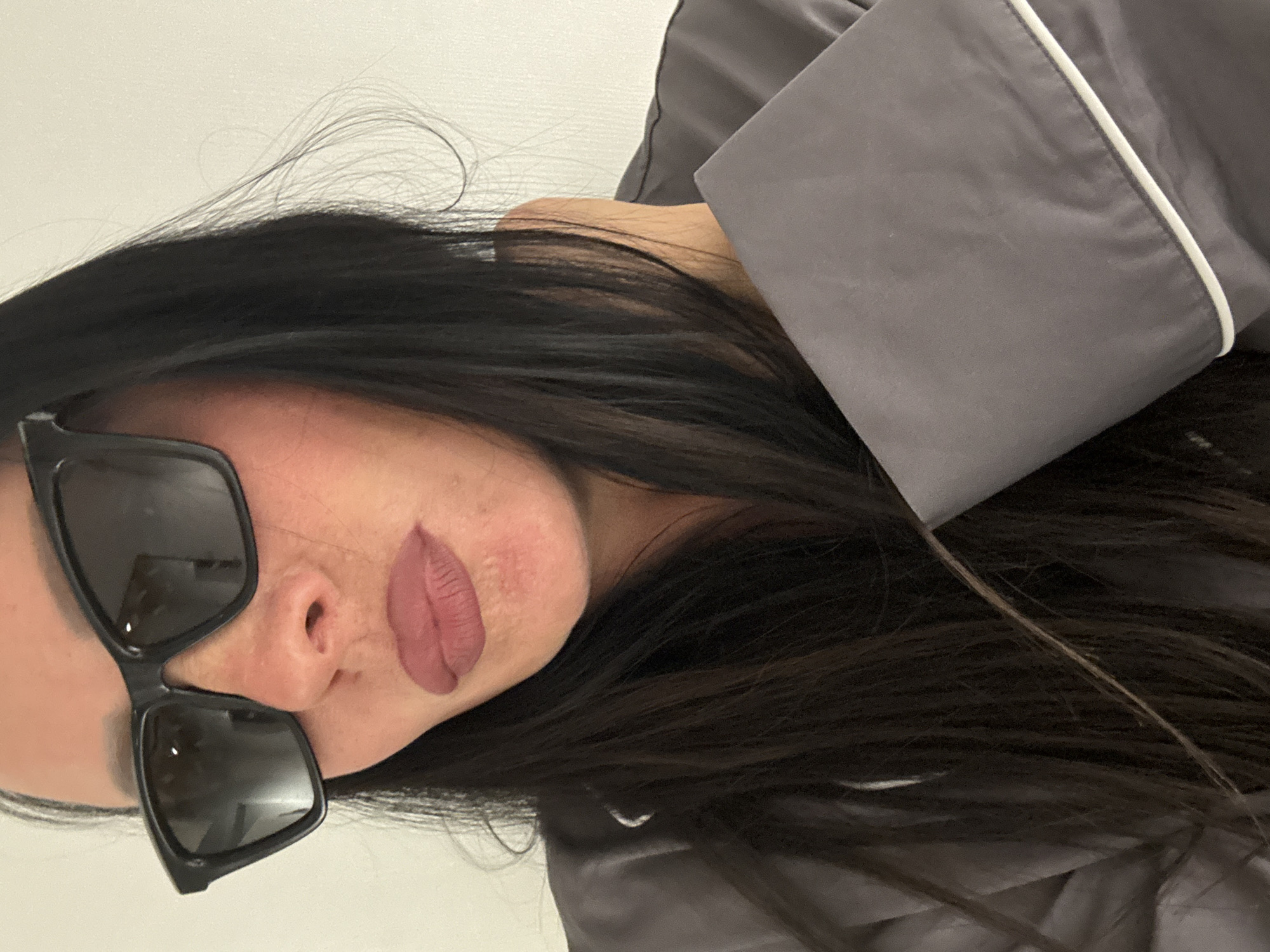 A woman with long, dark hair is wearing sunglasses and grey pajamas with white piping. The background appears to be a plain, white wall. There is no information to confirm this image was taken in Chaikovsky, Russia.