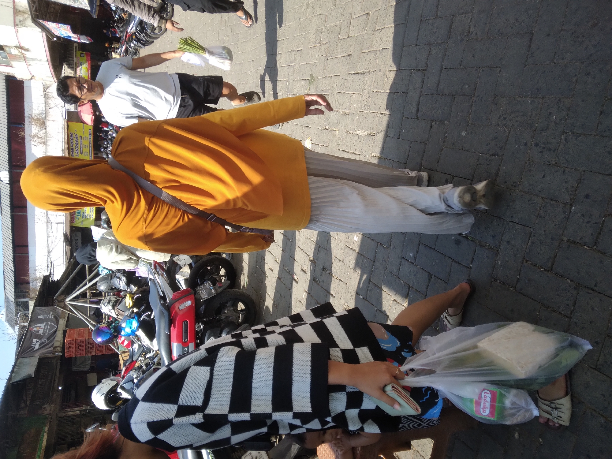 The image captures a street scene in Purwokerto, Indonesia, likely a market area, given the presence of parked motorcycles and stalls in the background. A person in the foreground from the back is wearing a bright orange hijab and loose-fitting, light-colored pants. They are walking away from the camera, with a dark-colored strap across their shoulder. To their left, another person is partially visible, wearing a black and white checkered top and carrying two plastic bags of groceries. One bag contains a green bottle labeled "Ladaku".
In the middle ground, a man wearing a light gray t-shirt and dark shorts is walking towards the right side of the frame, holding a bunch of green vegetables in a plastic bag. Several parked motorcycles are visible in the background, along with market stalls and buildings. The ground is paved with dark gray cobblestones, and the scene is brightly lit by sunlight, casting shadows on the ground. The overall impression is a busy, everyday scene in an Indonesian city.