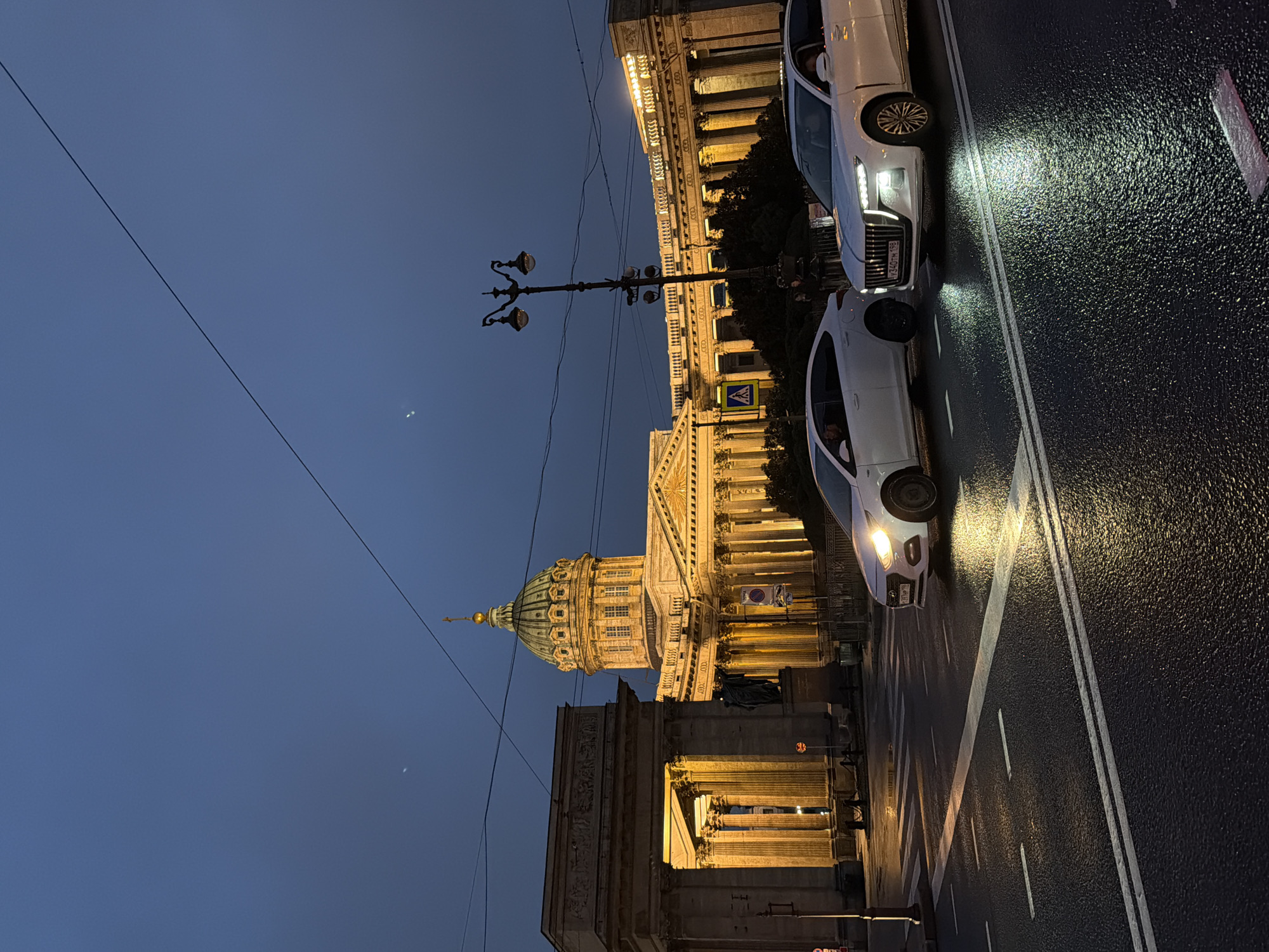 This is a nighttime, eye-level, medium shot of the Kazan Cathedral in Saint Petersburg, Russia. The sky is a deep, dark blue with faint hints of two celestial bodies, possibly the moon or planets. A thin, dark wire stretches diagonally across the upper portion of the frame. The illuminated Kazan Cathedral, with its iconic green dome topped by a golden cross, dominates the background. The cathedral is a neoclassical style building with a semicircular colonnade. To the left of the cathedral is another imposing structure with large columns and a classical facade, also illuminated. In the foreground, two white sedans are on a wet, dark asphalt road, with white lane markings. The headlights of both cars are on, casting bright beams onto the road, reflecting the wet surface. The car on the right has its license plate visible, reading "Y340TH 199". A street lamp with two globes stands between the cars and the cathedral, also illuminated. The overall atmosphere is one of urban twilight, with the grandeur of the architecture contrasted by the modern vehicles and wet street.