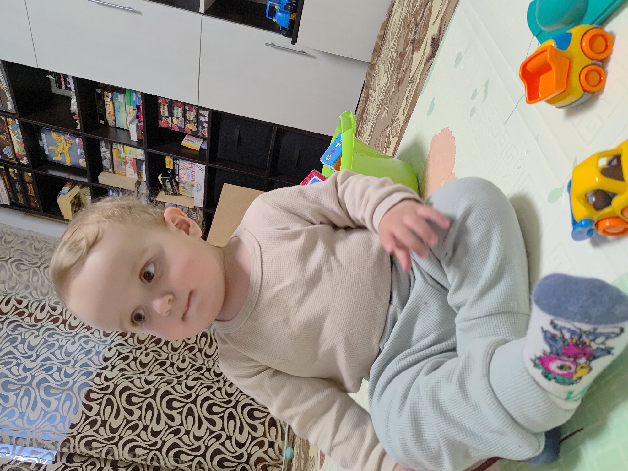 A young child, likely a toddler, is captured sitting on a light-colored play mat in an indoor setting. The child has light blonde hair, brown eyes, and is looking towards the camera with a neutral to slightly curious expression. They are dressed in a light beige long-sleeved waffle-knit shirt, light blue waffle-knit pants, and white socks with blue cuffs featuring a colorful, cartoon-like design on the ankle.
The scene depicts a domestic environment, possibly a child's play area or living room. Beneath the child, a patterned rug or blanket with swirling brown and light designs is visible. On the play mat to the child's right, several brightly colored plastic toys are present, including two orange and yellow toy trucks and a green plastic container or toy.
In the background, a white and dark brown shelving unit or cabinet holds various items. The dark brown shelves are filled with what appear to be books, board games, or video game cases. Visible text on one yellow box on a shelf reads "CARROMAN BARBARUS". Another white box on a lower shelf displays partial Cyrillic text that appears to be "КОРОТКИЙ НО РОВЫ". A blue toy truck is also seen on a shelf further up. The lighting suggests it is daytime. The child appears to be in a moment of quiet observation or play.