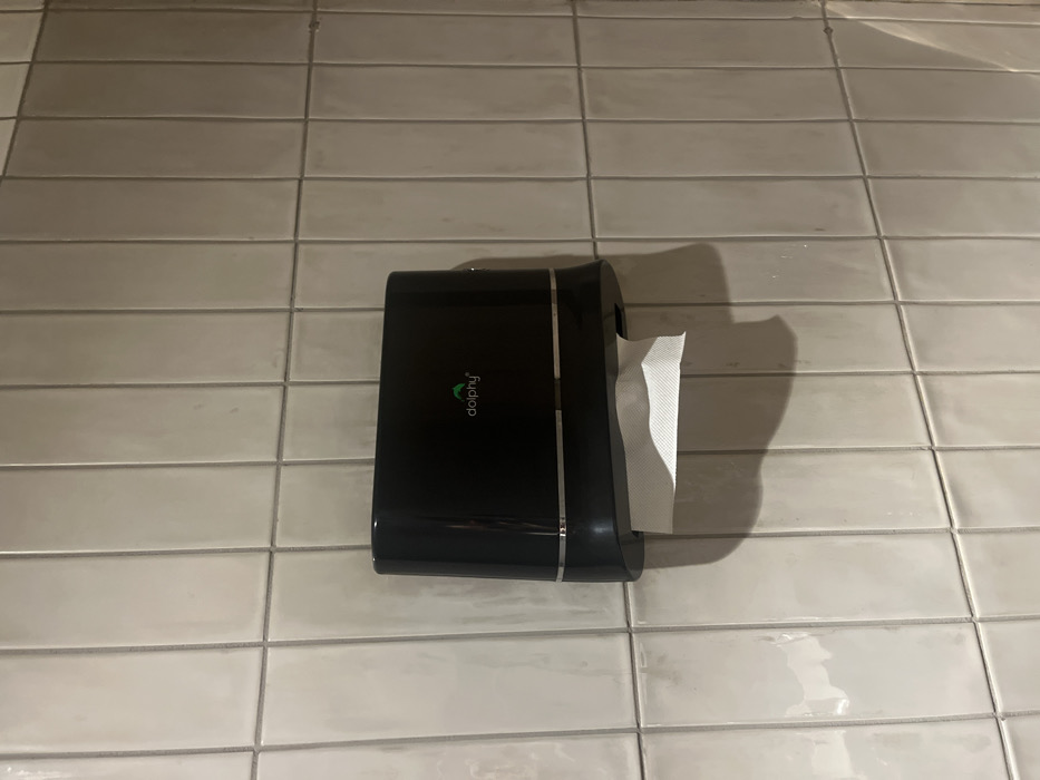 The image displays a black tissue paper dispenser mounted on a tiled wall. The dispenser is positioned centrally in the frame, slightly below the vertical midline. A white sheet of tissue paper is visible, partially dispensed from the right side of the holder.
The wall behind the dispenser is covered with light-colored, possibly off-white or beige, rectangular tiles. These tiles are arranged horizontally with dark grey grout lines, creating a grid pattern. The tiles have a subtle sheen, suggesting a glazed finish.
On the front surface of the black dispenser, the brand name "dolphy" is visible in a stylized green font, accompanied by a small green leaf or logo positioned above the letter 'd'. The dispenser casts a shadow on the tiled wall to its right, indicating an overhead light source.
The scene depicts an indoor setting, likely a restroom or kitchen in a public or commercial space in Dibrugarh, India, given the utility of such a dispenser and tiled walls. No activity is taking place; it is a static shot focused on the dispenser and its immediate environment. The lighting suggests a well-lit interior.