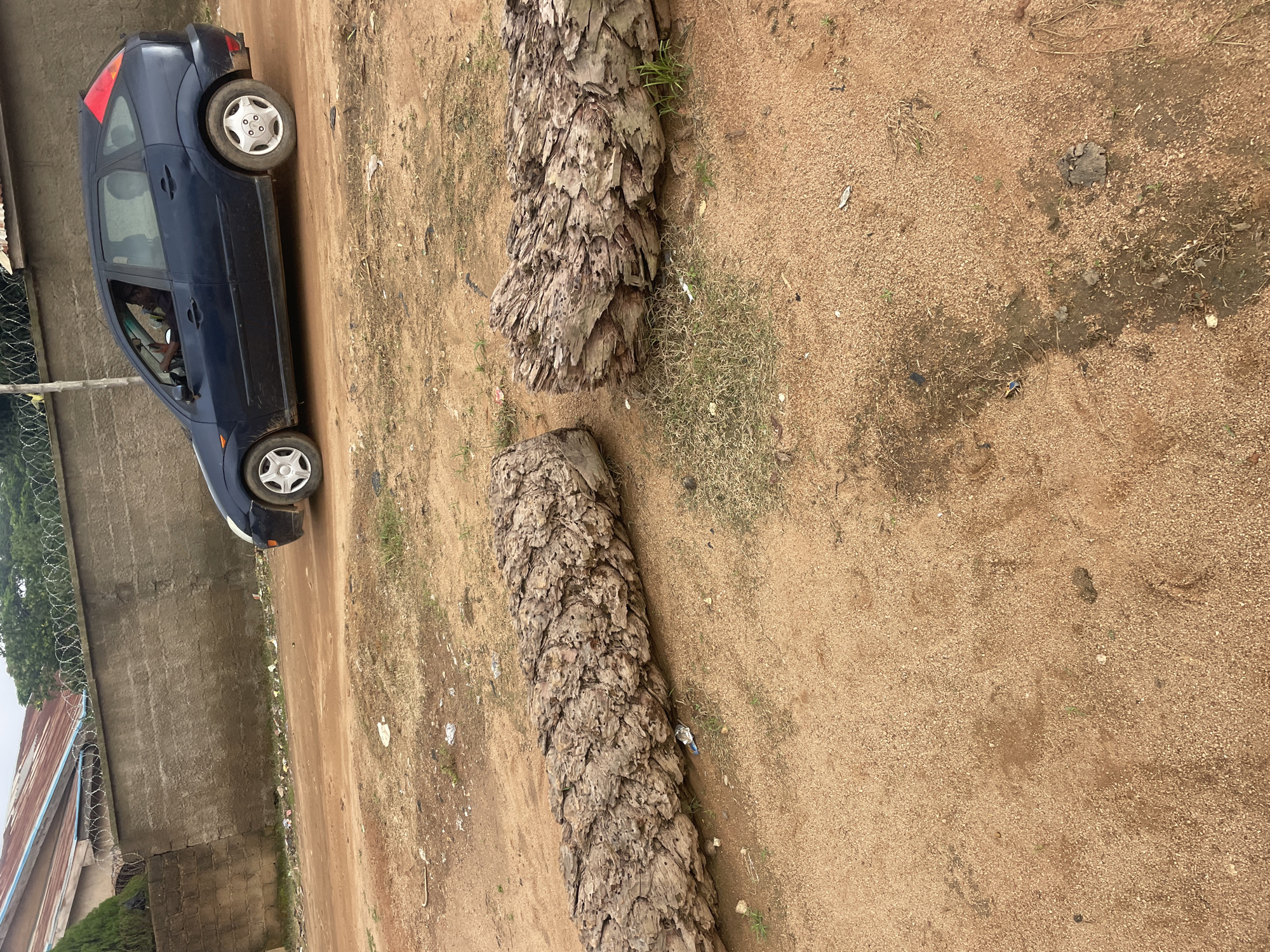 In Barakin Akawo, Nigeria, two large, weathered logs lie on a dirt road. A dark blue hatchback car with a red accent on the rear hatch drives away from the viewer on the same road. The car's driver is visible through the side window. A low, concrete wall topped with barbed wire runs along the background, separating the road from a building with a corrugated metal roof. The environment appears somewhat unkempt, with scattered debris and sparse vegetation.