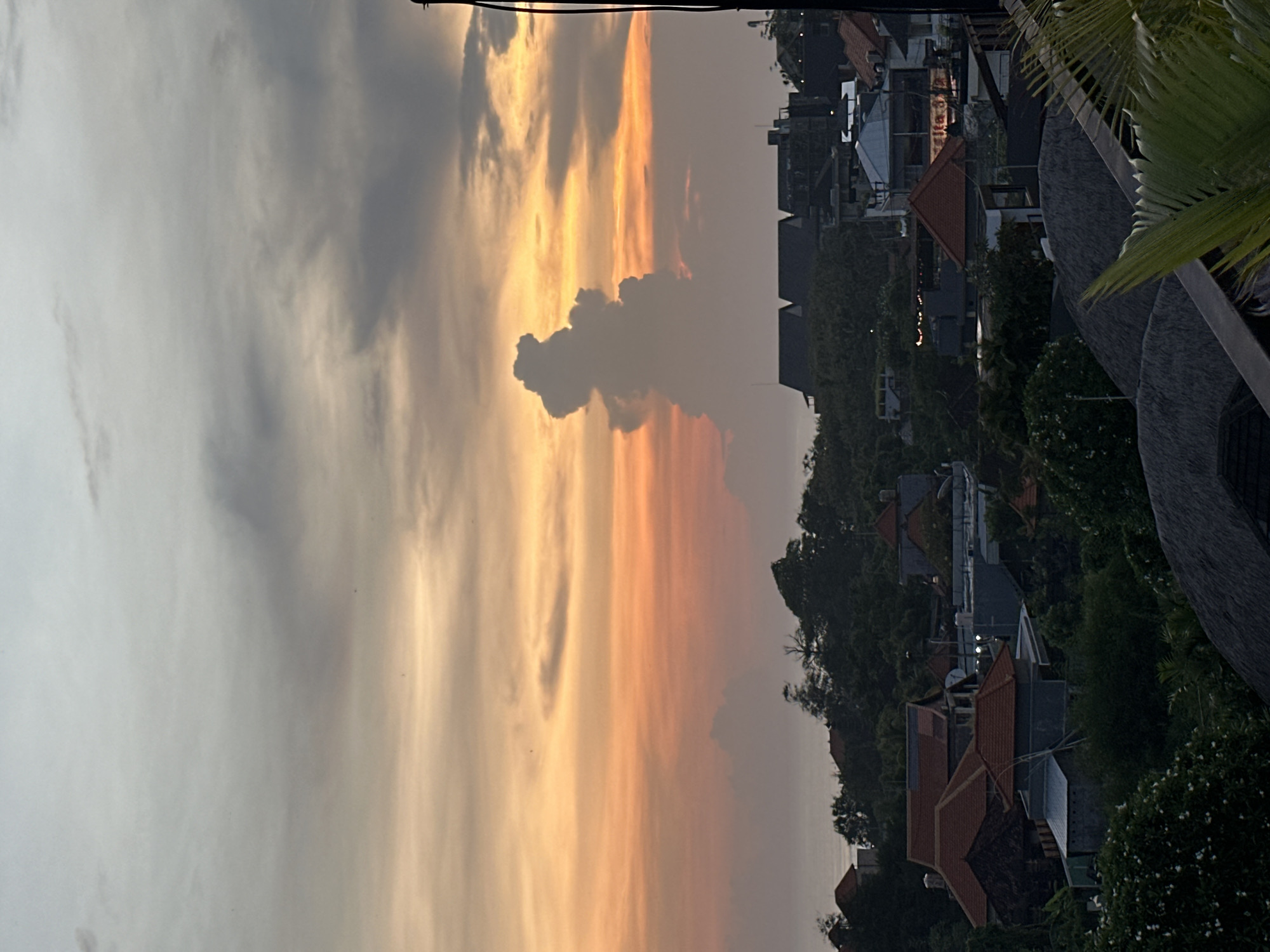 The image shows a sunset view from a building in Tibubeneng, Indonesia. The sky is filled with clouds, some dark and some illuminated by the setting sun in hues of orange and yellow. The silhouette of the rooflines and vegetation of the town are visible below the sky, adding depth to the scene. Some rooftops are visible along with dense tree foliage, and parts of a structure with thatched detailing are present in the foreground.