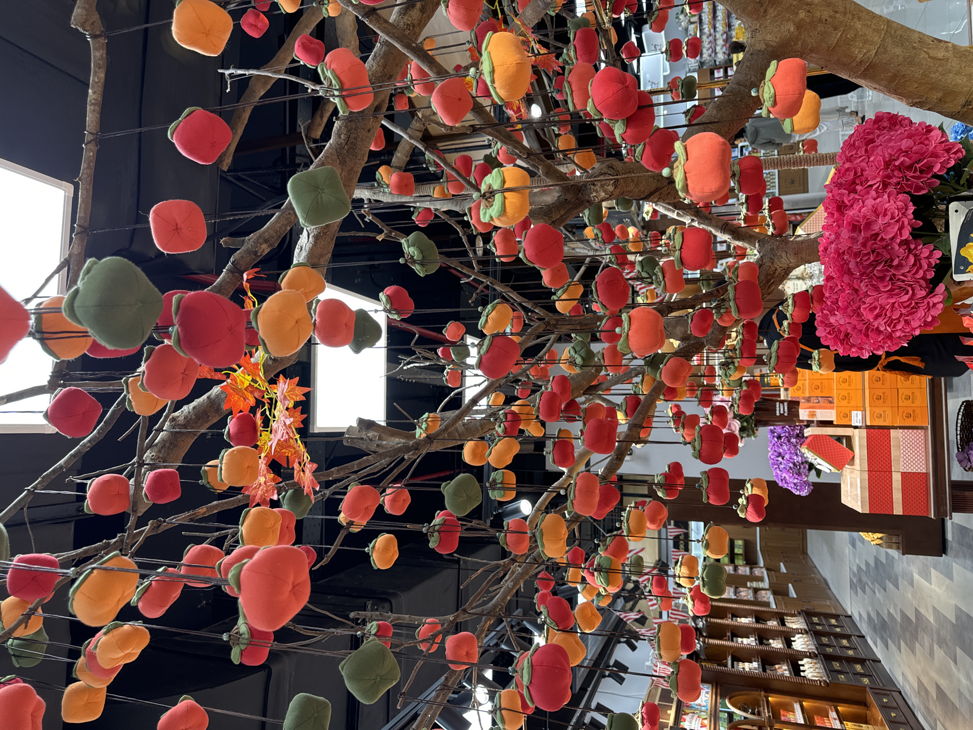 A low-angle, eye-level, indoor shot depicts an artificial tree decorated with fabric fruit hanging in the air. The tree has bare branches and is adorned with plush, fabric representations of persimmons or similar fruit in orange, red, and green hues. Some branches also feature artificial orange autumn leaves. The fruits appear to be suspended from the branches by strings or wires.
The background reveals a shop interior. Shelves stocked with various goods line the walls. The flooring has a grey and white geometric pattern. In the upper right corner of the image, a cluster of bright pink flowers adds a pop of color. The scene is well-lit with artificial lighting, and the overall impression is one of an artistic installation or decoration within a retail environment.
The location is Phu Thuy Ward, Vietnam.