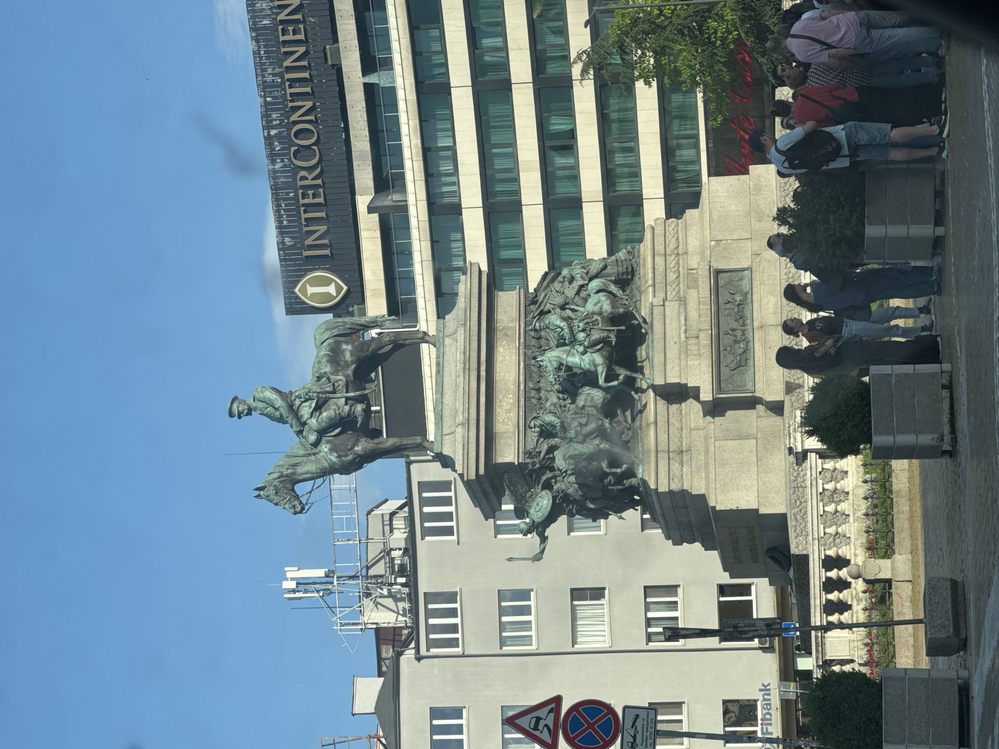 A bright, sunny outdoor scene captures a prominent bronze equestrian monument in a city setting. The central feature is a large, dark bronze equestrian statue of a figure, likely a military leader or monarch, on horseback, resting atop a grand stone pedestal. Below the main rider, the monument's base is adorned with intricate bronze reliefs depicting multiple human figures engaged in dynamic scenes, possibly historical battles or allegorical representations. The stone pedestal, made of light-colored material, also bears unreadable inscriptions.
Behind the monument, a tall, modern building features prominently. Its upper section displays a dark facade with the distinct gold lettering "INTERCONTINENTAL" and the brand's "I" logo. Below, the building's architecture is characterized by alternating light-colored vertical panels and dark-tinted windows. To the left of the monument's base, a multi-story, light grey building with numerous rectangular windows is visible, with scaffolding suggesting ongoing work on its upper floors. A "FiBank" sign can be seen near its base.
To the right of the monument, a group of people stands on a paved sidewalk, appearing to observe the surroundings or pass through the area. They are dressed in casual summer clothing. Trees and shrubs are interspersed around the monument and along the pedestrian zone, adding greenery to the urban landscape.
The sky is a clear, bright blue with minimal, wispy clouds, indicating fair weather during the day. At the bottom left, several traffic signs are present, including a triangular warning sign and a circular "no stopping" sign, along with other street furniture. The scene captures a vibrant public space in Sofia, Bulgaria, blending historical architecture with contemporary city life.