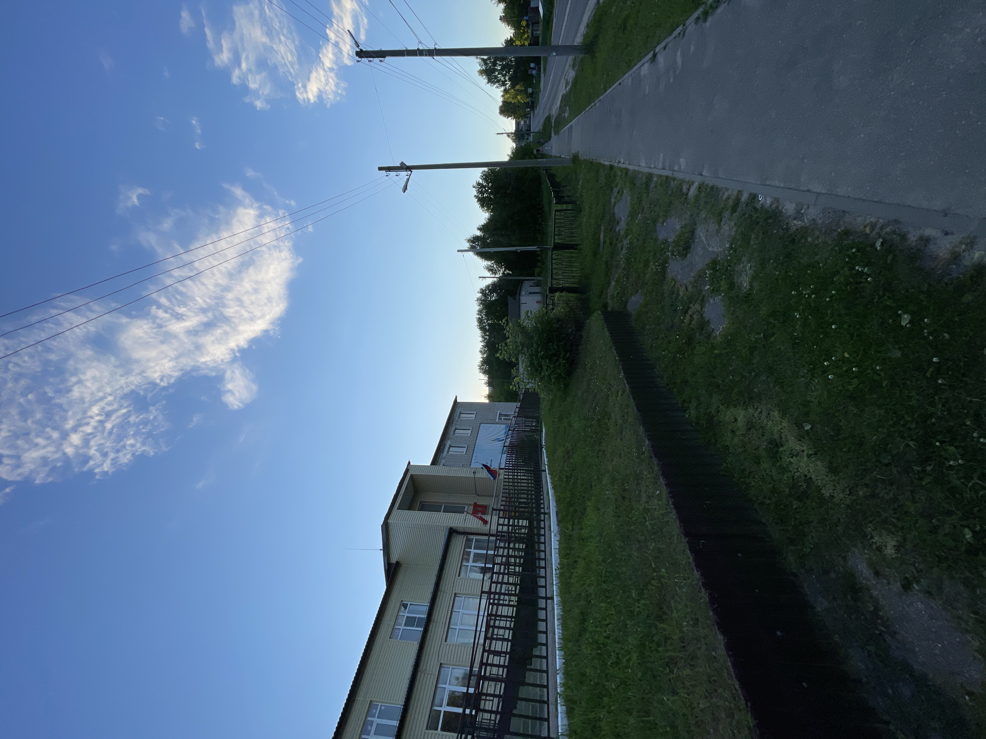 This is a vertical, eye-level, full shot of a building in Krasnye Baki, Russia, on a clear, late afternoon or early evening. The building, which appears to be a school or administrative building, is two stories tall and clad in light beige siding. It has a dark brown sloped roof and numerous windows. A metal ramp leads to the main entrance, with a railing. Two flags, one red and white, and another blue, white, and red, are displayed on the facade. A large blue banner with white lettering is visible on the right side of the building.
In front of the building is a grassy area, separated from a paved sidewalk by a low, dark, horizontal border. The sidewalk runs along the right side of the image, parallel to a road that is visible in the distance. Several utility poles with power lines are scattered along the sidewalk and road. The sky is a clear blue, with a few wispy white clouds. The overall atmosphere is calm and serene, with the late afternoon sun casting a soft light. The image does not contain any people or any apparent activity.