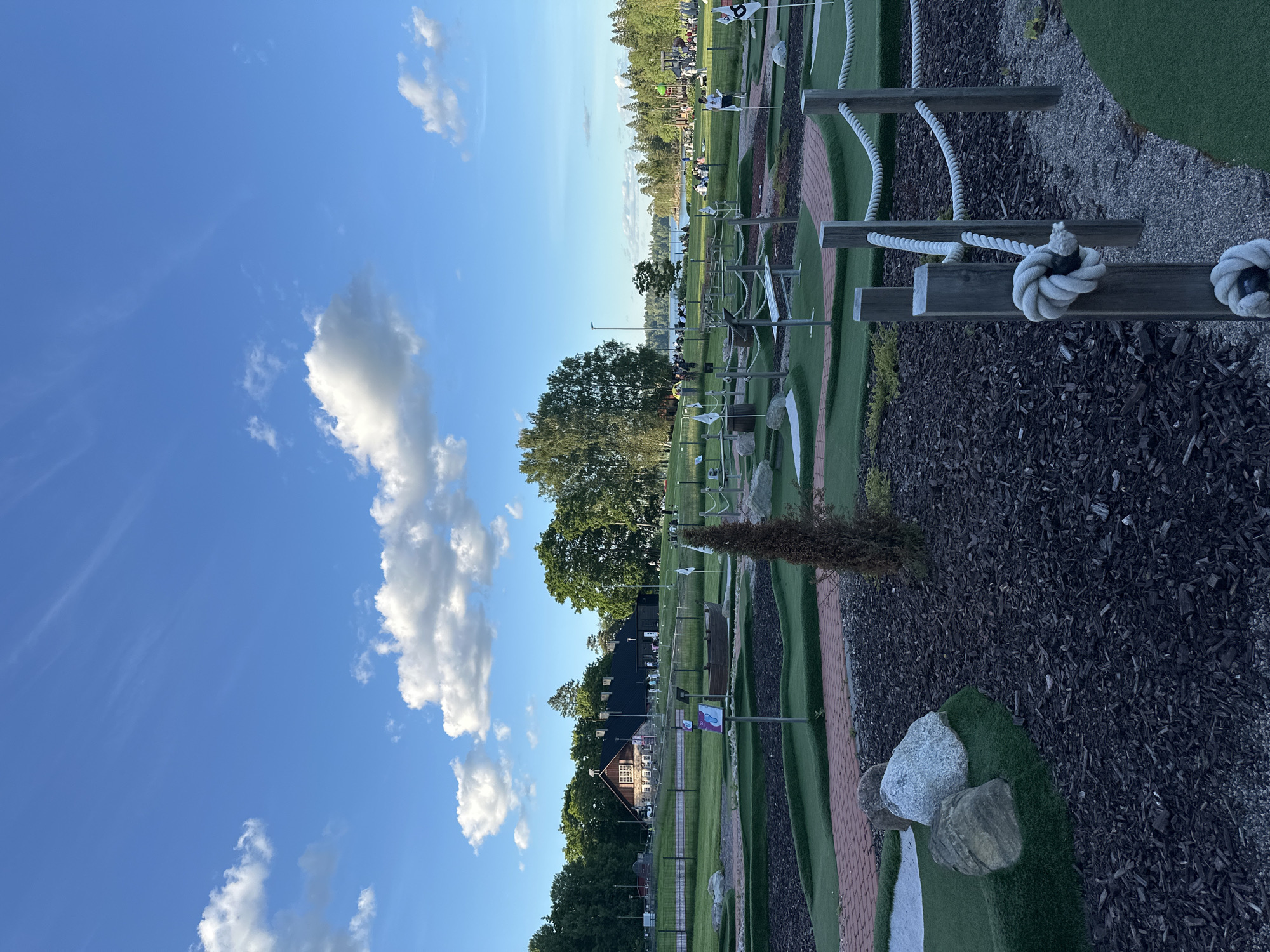 The image depicts a sunny outdoor scene at a miniature golf course with a body of water in the background. The foreground features a section of the miniature golf course with artificial turf, brick pathways, and decorative rocks. Wooden posts with rope railings are visible in the right foreground, suggesting a boundary or a decorative element. In the midground, more of the miniature golf course is visible, including several holes with flags. Beyond the golf course, a large building with a dark roof and a stone facade is situated amongst trees. Several people are scattered around the building and on the grounds, some appearing to be patrons or staff. Further in the background, a body of water, likely a lake, is visible, with trees and a treeline on the opposite shore. The sky is blue with scattered white clouds, indicating fair weather.