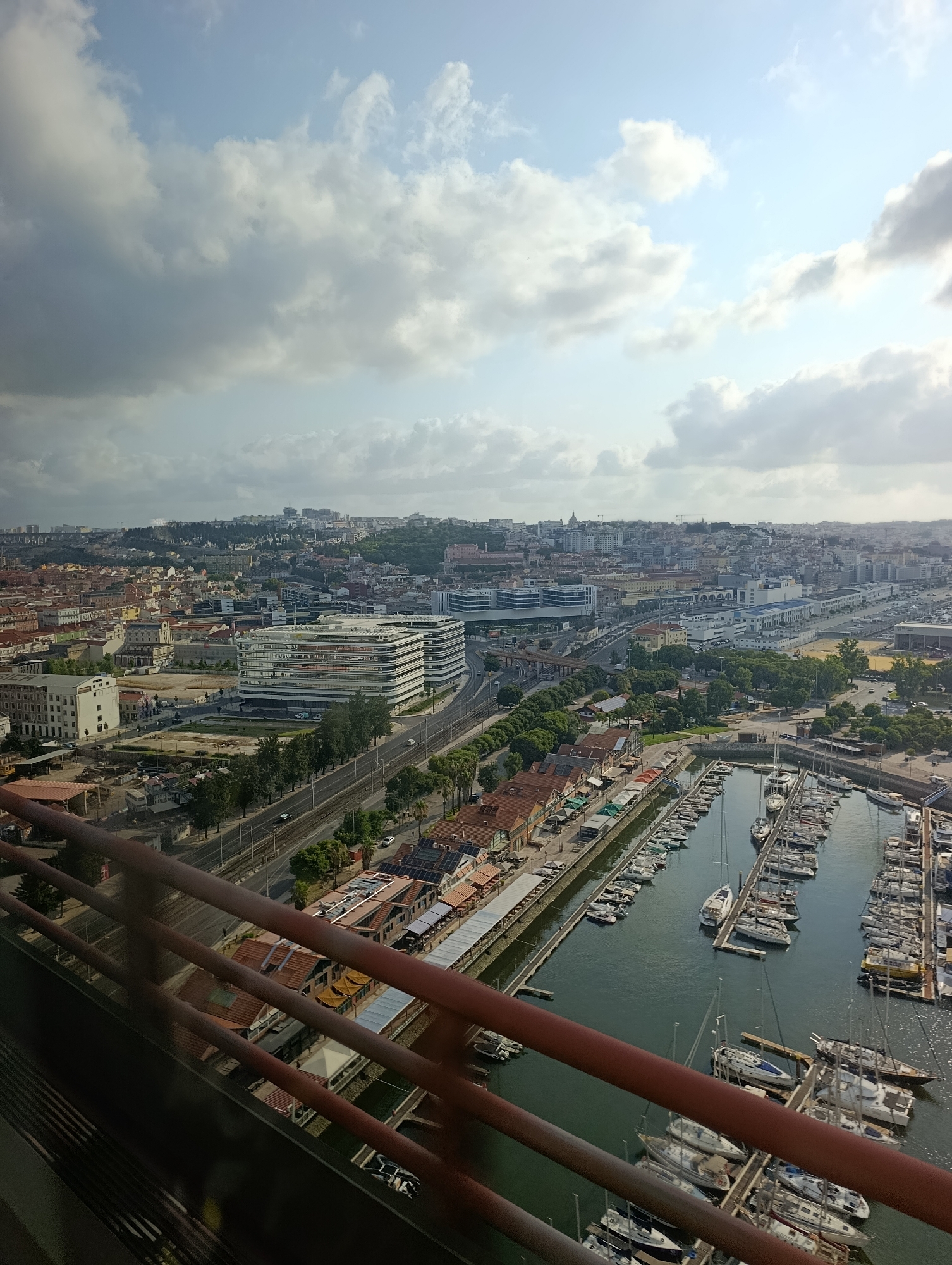 This is an aerial, high-angle shot of a city harbor in Lisbon, Portugal, on a partly cloudy day. In the foreground, a red metal railing is visible, obscuring a portion of the view. The harbor is filled with numerous docked sailboats and yachts of varying sizes. Beyond the harbor, the cityscape unfolds with a mix of residential buildings with terracotta roofs and modern office buildings with glass facades. A tree-lined street runs parallel to the harbor, with a few cars visible. In the distance, the city extends across rolling hills, with more buildings and structures scattered throughout. The sky is a mix of blue sky and scattered white clouds.