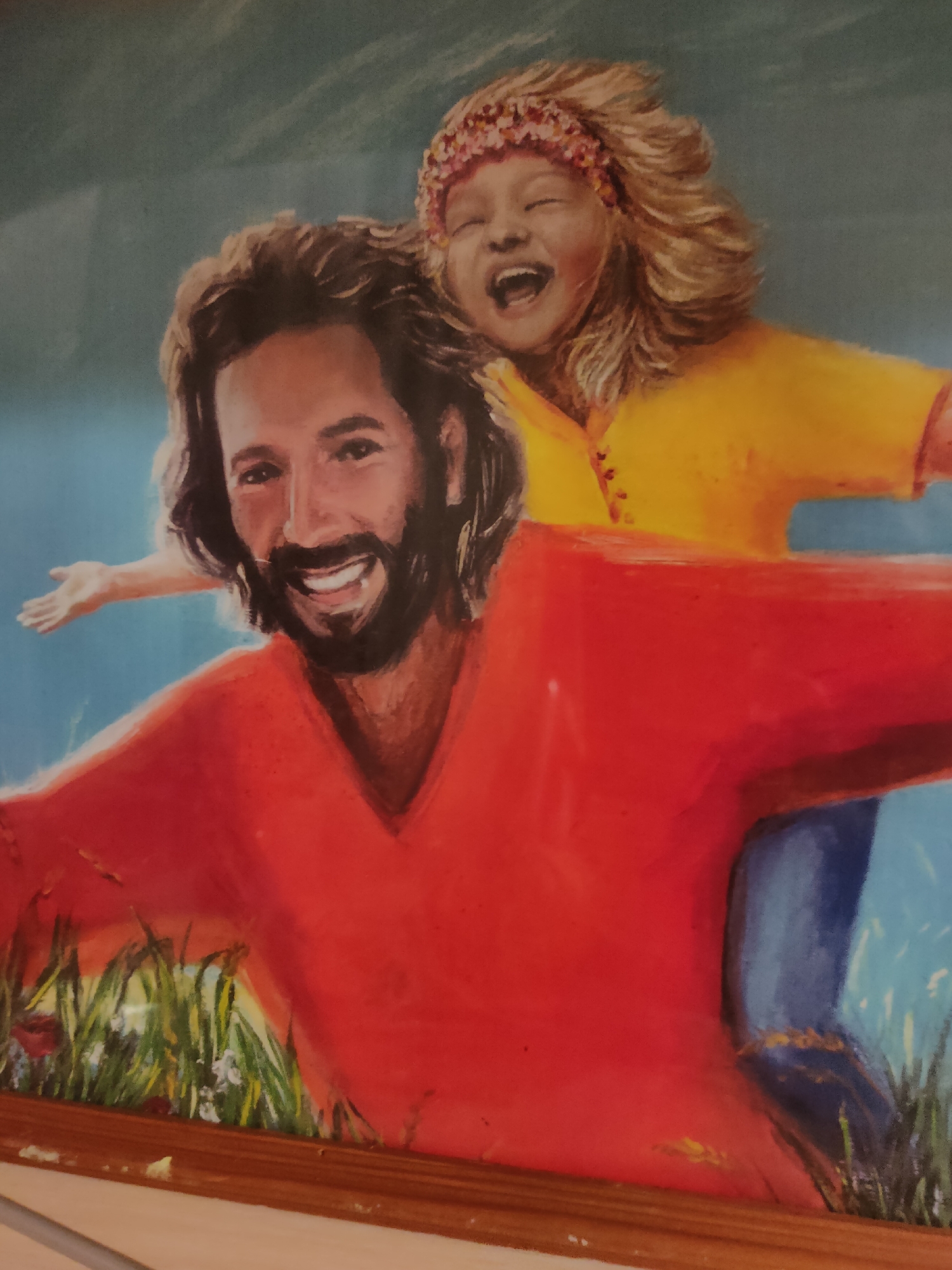 This image is a painting of a man and a child. The man has long brown hair and a beard, and is wearing a red shirt. He is smiling and has his arms outstretched. The child is riding on the man's back and is wearing a yellow shirt and a pink headband. The child is laughing and has their arms around the man's neck. They are in a grassy area with a blue sky in the background. The painting appears to be in a home or a public building in Oktyabrsky, Russia.