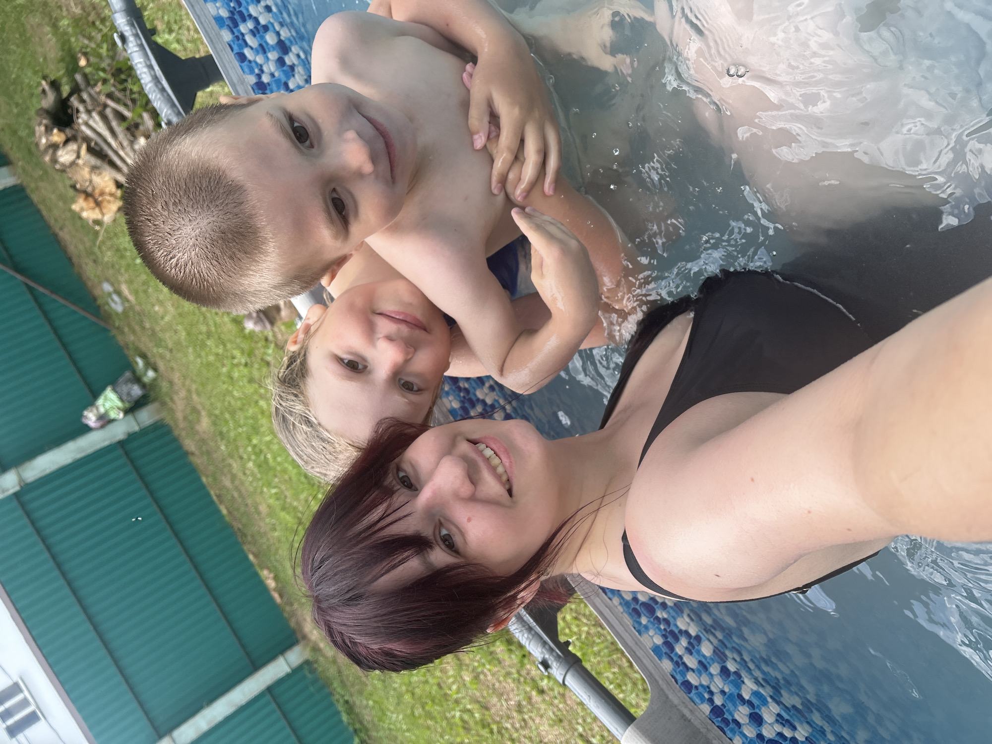 In the city of Fokino, Russia, a woman with dark hair and a black swimsuit smiles at the camera while taking a selfie in an outdoor inflatable pool. She is holding two young boys, one of whom is embracing her. The pool is filled with clear water and has a blue and white tiled pattern on the inside. Behind the pool, a green wooden fence encloses a grassy area. There is a pile of cut wood near the fence, and a building can be seen in the background. The overall scene depicts a family enjoying a summer day.