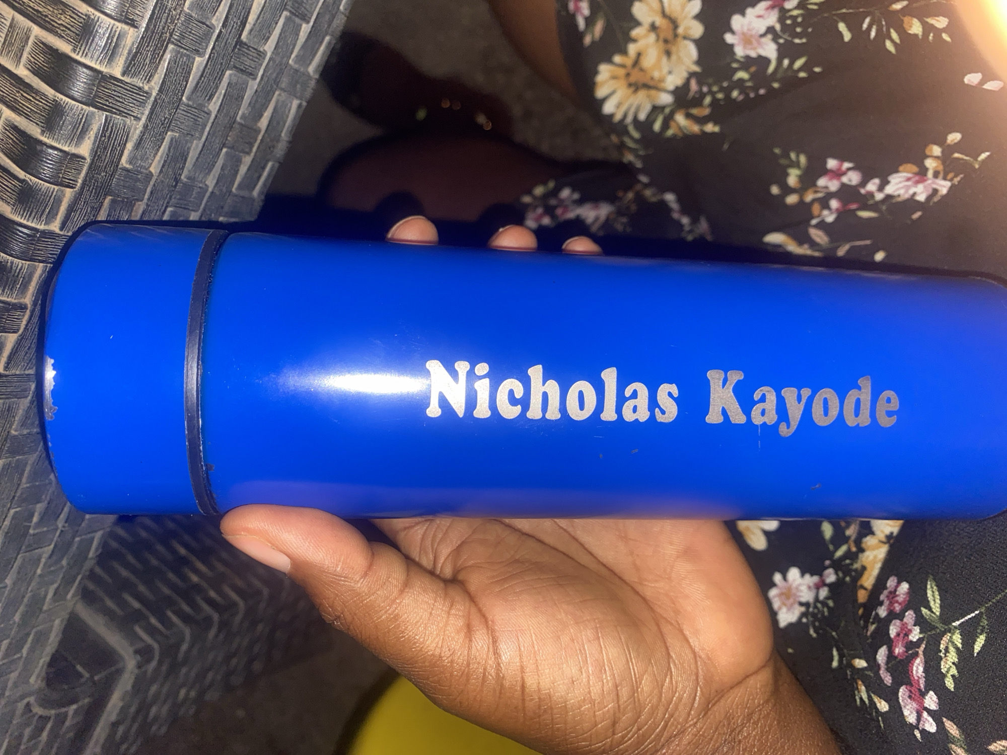 A blue water bottle with the name "Nicholas Kayode" engraved in silver lettering is held in a person's hand. The person's arm is visible, and they are wearing a black floral dress. To the left of the bottle is a gray wicker chair. It is not possible to verify if the image was taken in Television, Nigeria.