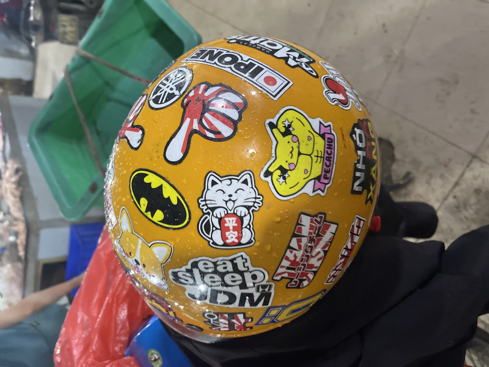 This close-up, high-angle image showcases a bright yellow motorcycle helmet heavily decorated with numerous stickers, all covered in small water droplets. The helmet appears to be resting, possibly on a motorcycle seat or stand, within a casual setting in Bac Nha Trang Ward, Vietnam.
The stickers on the helmet are a blend of pop culture, automotive, and Japanese themes. Notable decals include a small rectangular Japanese flag and the text "IPONE." A circular sticker displays the "YAMAHA" logo, while another features a stylized fist with a Japanese rising sun pattern. A humorous "PECACHU" (likely Pikachu) sticker depicts a muscular yellow character. The iconic black "Batman" logo is also present.
Further down, a white Maneki-neko (waving cat) sticker is visible, holding a red sign with the Japanese characters "平安" (Heian), which translates to peace or tranquility. Nearby, a cute, cartoonish Corgi dog sticker adds to the playful customization. The phrase "eat sleep JDM" is prominently displayed, referencing Japanese Domestic Market car culture. Several other stickers feature various Japanese text and stylized designs, though not all are fully decipherable.
The small water droplets covering the helmet indicate that it has recently been exposed to rain or a humid environment. In the blurred background, parts of a person's arm and leg are visible, along with a green plastic crate and a red plastic bag, all resting on a light-colored tiled floor, common in Southeast Asian outdoor or semi-outdoor spaces. The image highlights a vibrant and personalized piece of riding gear.