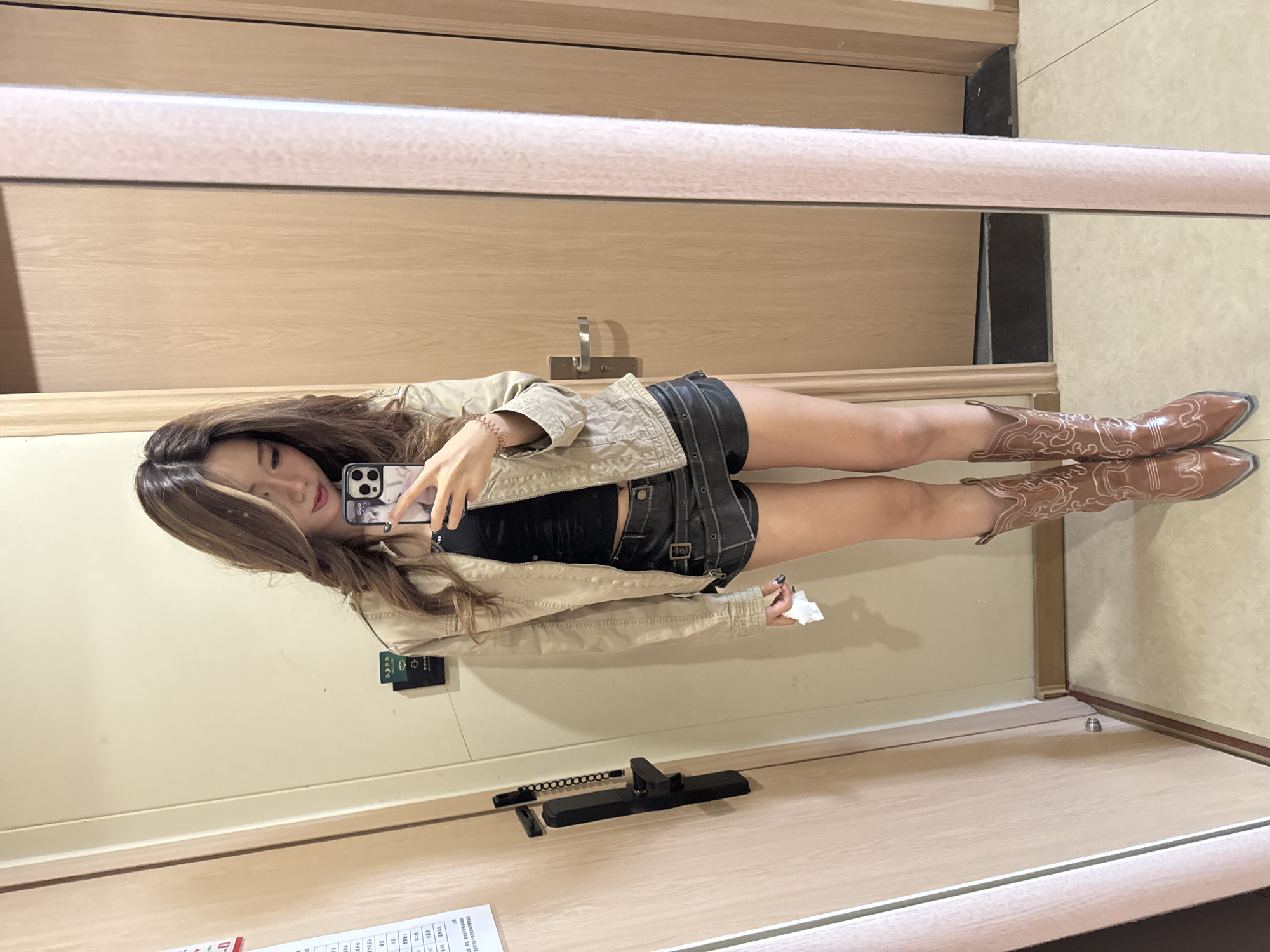 A woman in a mirror selfie, wearing a tan jacket, black top, short black skirt, and cowboy boots. She is holding a smartphone, with a partial view of a room in the background. The mirror reflects light-colored wooden panels and a portion of a light-colored floor. A hotel room key card appears in the mirror. There are Chinese character documents on the lower left corner of the image. The setting suggests a hotel room in Dunhuang City, China.