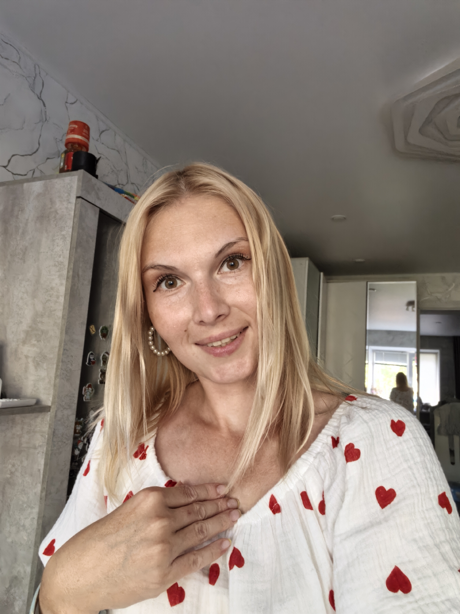 A medium shot captures a blonde woman with fair skin and freckles looking towards the camera with a slight smile. She has brown eyes and light makeup, including eyeliner and mascara. She is wearing a white blouse with a pattern of small red hearts. Her right hand is placed on her chest. She has long, straight blonde hair.
The background suggests an indoor setting, possibly a kitchen or living area. To the left, a gray cabinet is visible with a red jar on top. The wall behind it has a decorative marbled pattern. To the right, a large mirror reflects another woman standing further back in the room. The ceiling features an interesting architectural detail. The scene appears to be a personal photograph taken within a residential space. The image is taken from a slightly low angle.
