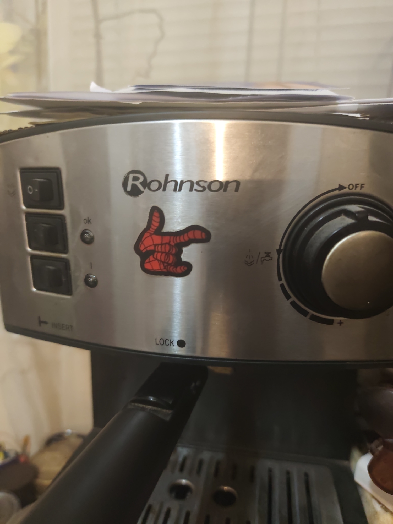 A close-up shot focuses on the top and front of a stainless steel Rohnson coffee maker. A stack of papers is precariously balanced on top of the appliance. On the control panel, there are three black toggle switches on the left, the top one in the "0" (off) position. To the right of the switches, there are two small indicator lights, one labeled "ok" and the other with a symbol that is difficult to discern. A red sticker of a hand with outstretched fingers, resembling a superhero gesture, is positioned to the right of the lights. Further to the right, a large black dial with a metallic gold center is used for controlling settings, with markings indicating "OFF" and "+" for increased intensity, and icons for steam and coffee. Below the dial, a symbol for "LOCK" is visible. The overall environment appears to be indoors, with a neutral-toned, textured wall in the background. The lighting suggests it is daytime.