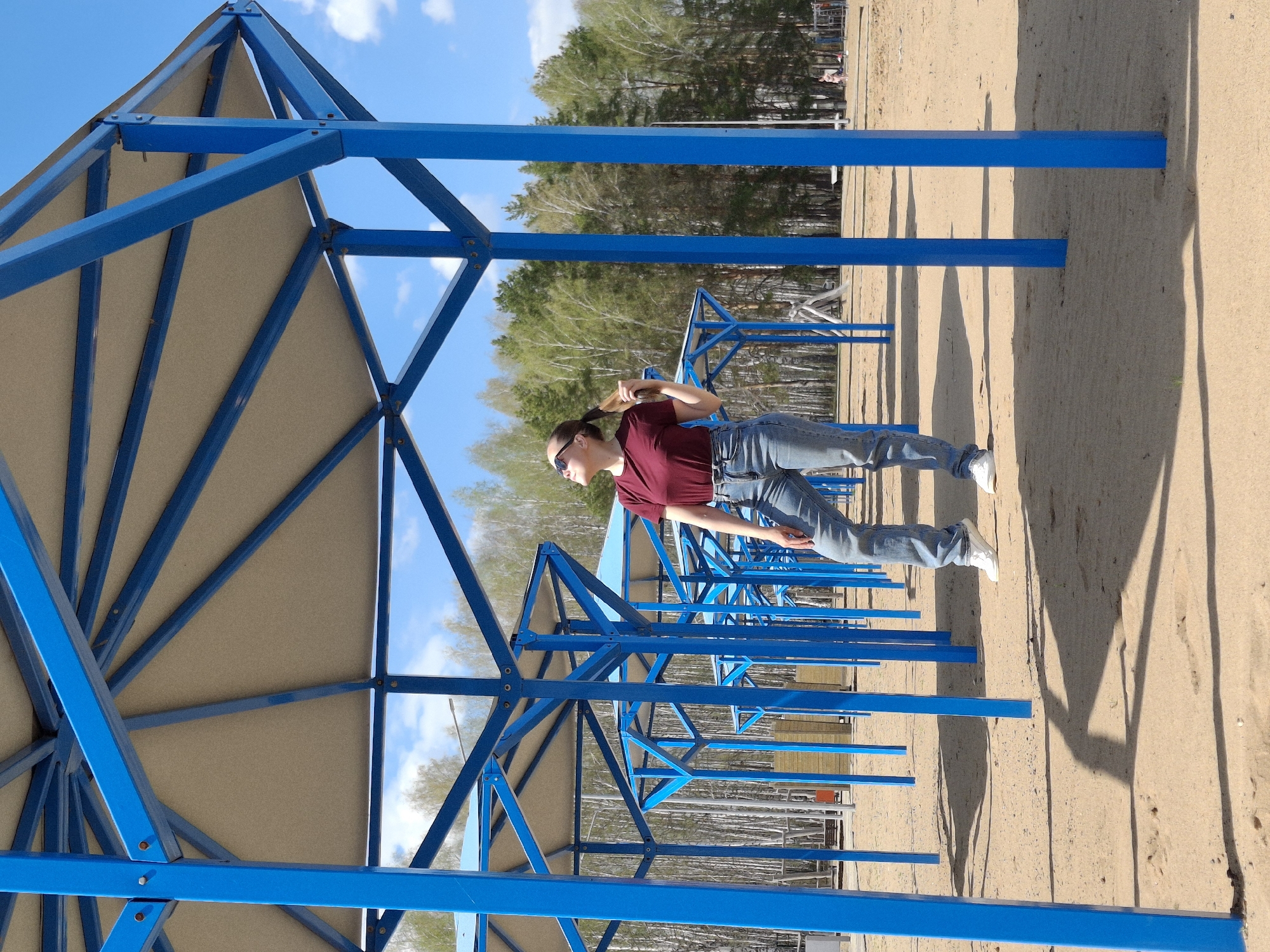 A full-length portrait captures a woman from a side perspective on a beach in Chaikovsky, Russia. She is seen in a maroon short-sleeve shirt and jeans, with her hair pulled back. She is standing under a series of identical blue metal structures with tan-colored roofs that provide shade. The ground is sandy. In the background is a line of trees. The sky is visible in the upper part of the frame with some scattered clouds.