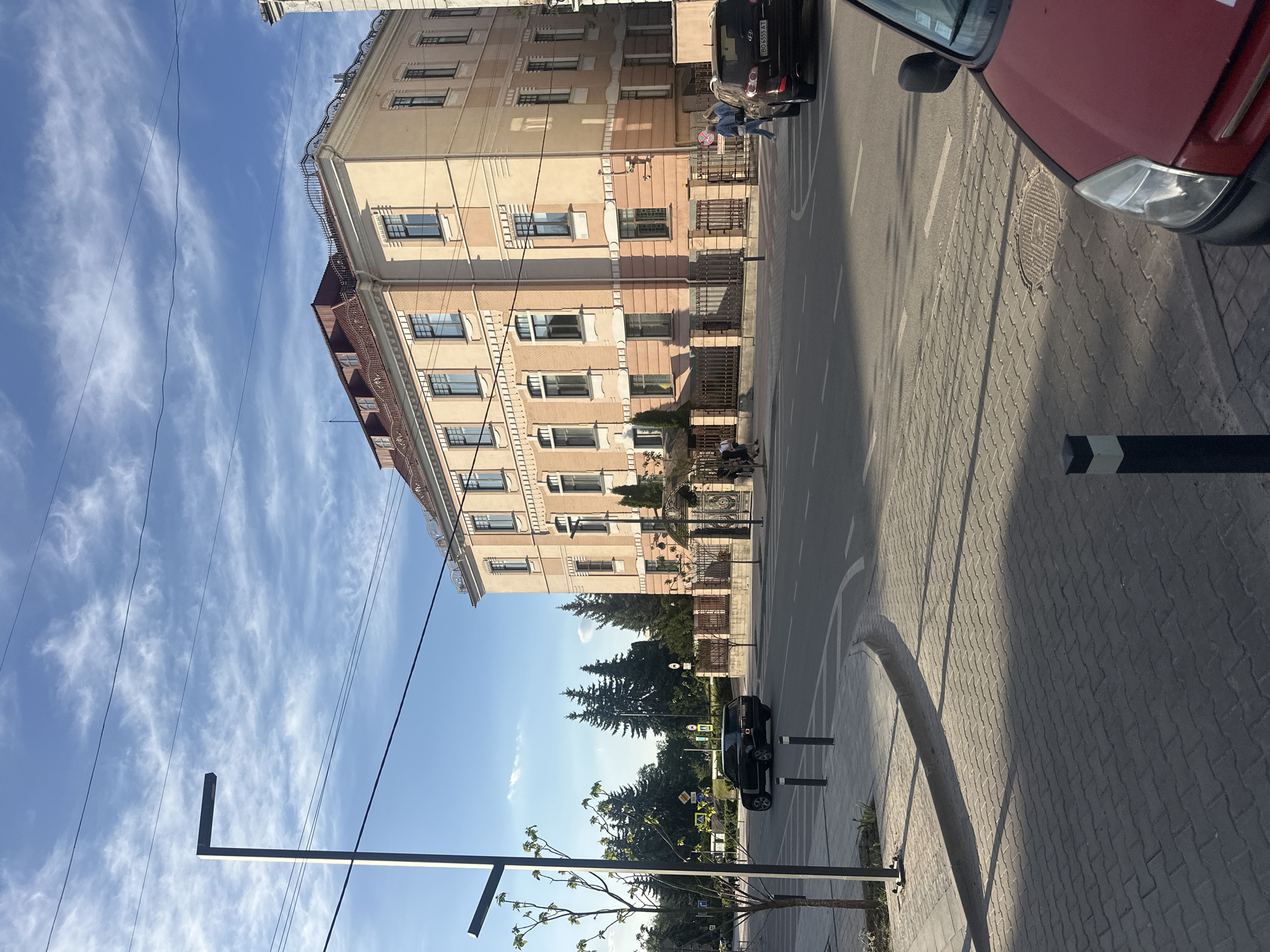The image depicts a street scene in Ternopil, Ukraine, under a blue sky with wispy clouds. A multi-story building with a light orange facade dominates the left side of the frame, displaying multiple windows and detailed architectural elements. Adjacent to the building is a paved road with white lane markings and a sidewalk constructed of interlocking pavers. Several trees line the sidewalk. A few vehicles are present, including a red car partially visible on the right side of the frame and a black car parked along the street. Two individuals are captured in motion near the building, while another person is seen walking on the sidewalk in the distance. Various street signs and light poles are visible.