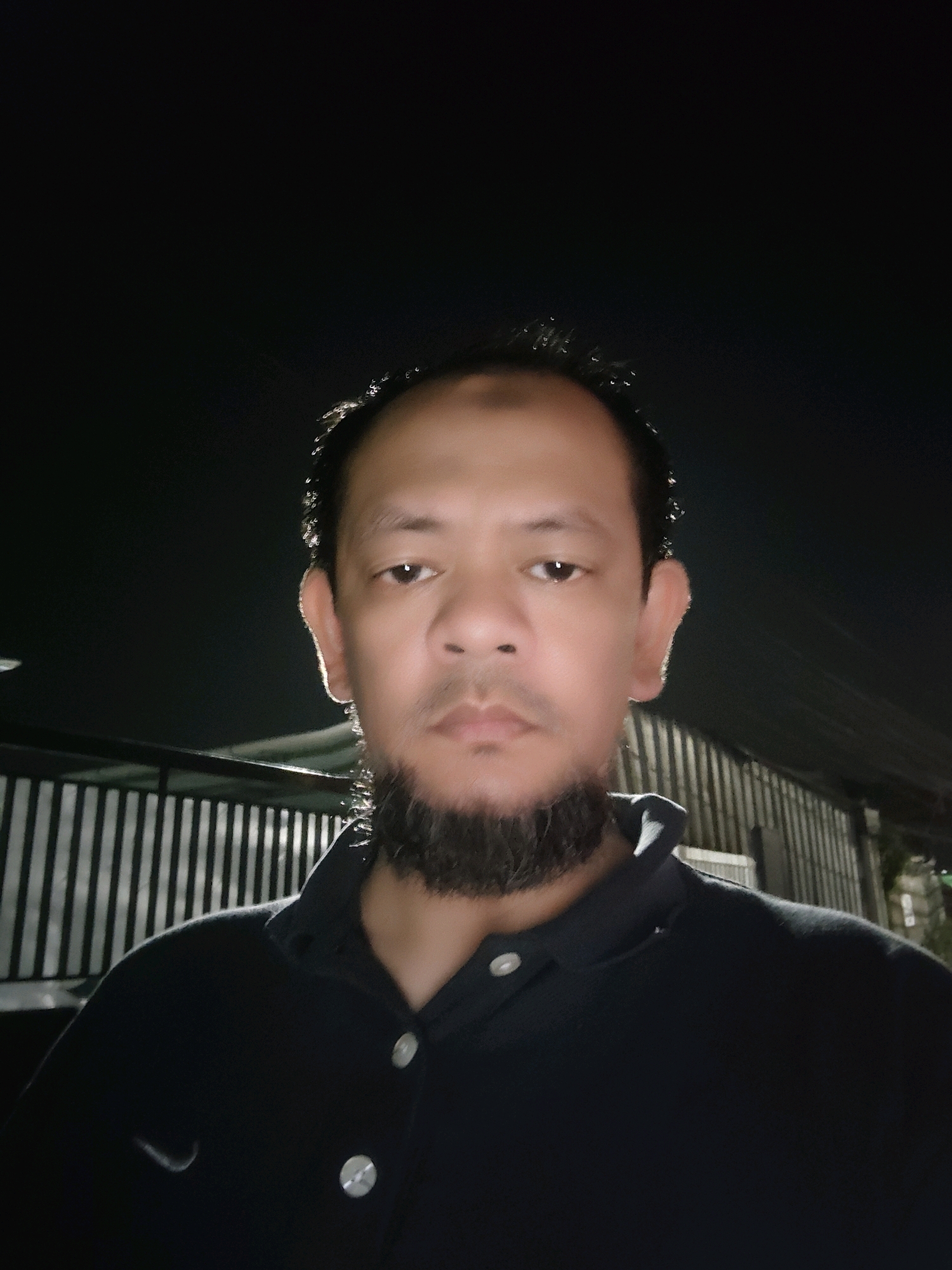 A medium shot shows a man with dark hair and a beard, wearing a dark polo shirt, standing outside at night. He is looking directly at the camera. Behind him, a fence made of vertical metal bars is visible, separating him from a corrugated metal structure. The background is dark, suggesting it is nighttime in an outdoor urban setting, possibly within the city of Bogor, Indonesia.