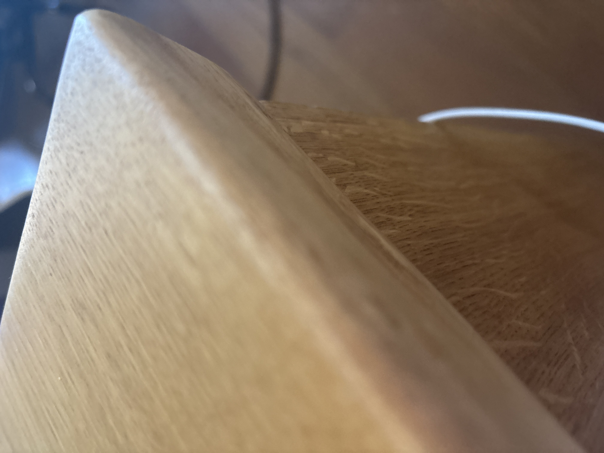 The image is a close-up, eye-level, medium shot of the corner of a light-colored wooden surface, likely a table or countertop. The wood grain is visible, running diagonally across the frame from the upper left to the lower right. The surface has a rounded edge. The background is out of focus and appears to be a wooden floor and possibly some dark objects. A thin white cord or wire is visible in the lower right quadrant, curving upwards along the side of the wooden surface. There are no people or animals visible in the image. The scene appears to be indoors. The location is not discernible from the image itself, therefore no specific details about the city of Orekhovka, Russia can be provided.