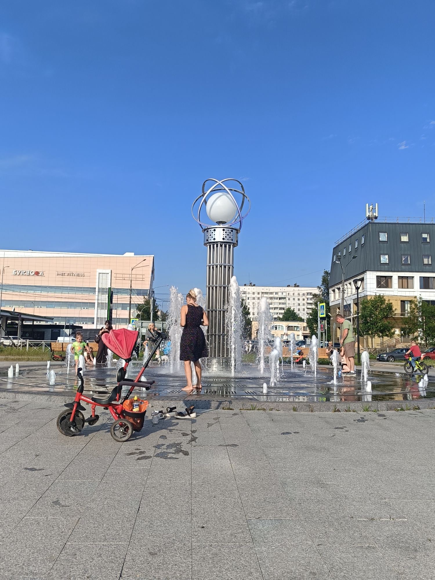 A wide-angle, eye-level shot captures a bright, sunny day in an outdoor public plaza featuring a fountain. The plaza is paved with large, grey granite tiles, with some patches of darker, wet stone indicating the spray from the fountain. In the background, a modern, multi-story building with a dark grey, angled roof and white accents dominates the right side of the frame, adorned with rooftop equipment. To the left, a lighter pink, rectangular building with prominent signage, including "Fit Fitness" and Cyrillic text, stretches across the mid-ground, bordered by a bus stop and a grassy strip.
In the foreground, a red and black child's tricycle is parked on the plaza tiles. Several individuals are interacting with the fountain, which features numerous vertical jets of water creating a refreshing atmosphere. A woman in a dark, polka-dot dress stands barefoot in the shallow water of the fountain, her back to the camera. Nearby, a young girl in a green outfit is running through the water, her feet splashing. Another child, partially visible, is also playing in the fountain. Two adults stand near the edge of the fountain, one a woman in dark clothing and the other a man in a green t-shirt and shorts, observing the children. Further back, a person on a yellow bicycle with a red helmet rides through the plaza. The sky is a clear, vibrant blue. The scene suggests a recreational area within a city, likely a park or a pedestrian zone, where people are enjoying the warm weather and the water feature. The presence of the buildings and the asphalt tiling indicate an urban setting.