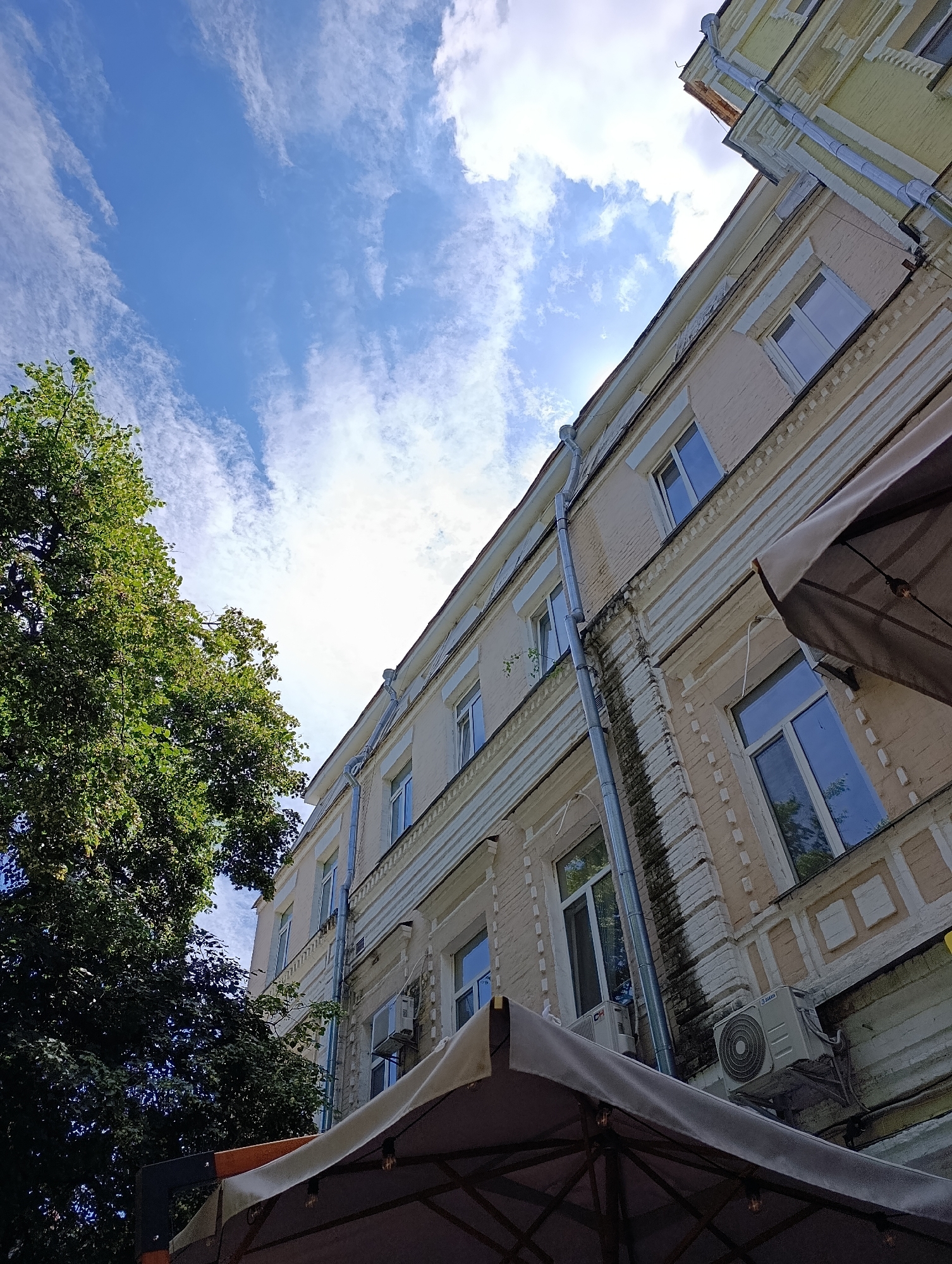 This is a low-angle, upward-facing, outdoor shot of a building facade in Kyiv, Ukraine, with a tree on the left and a large umbrella in the foreground. The building is old, with a light beige brick facade and ornate white decorative trim around the windows and along the horizontal lines between floors. Several windows with white frames are visible, reflecting the blue sky and white clouds. A metal downspout runs vertically along the side of the building. The tree on the left is lush and green. The umbrella in the foreground is a dark color, possibly brown or gray, and obscures the lower portion of the building. The sky is a bright blue with scattered white, wispy clouds.