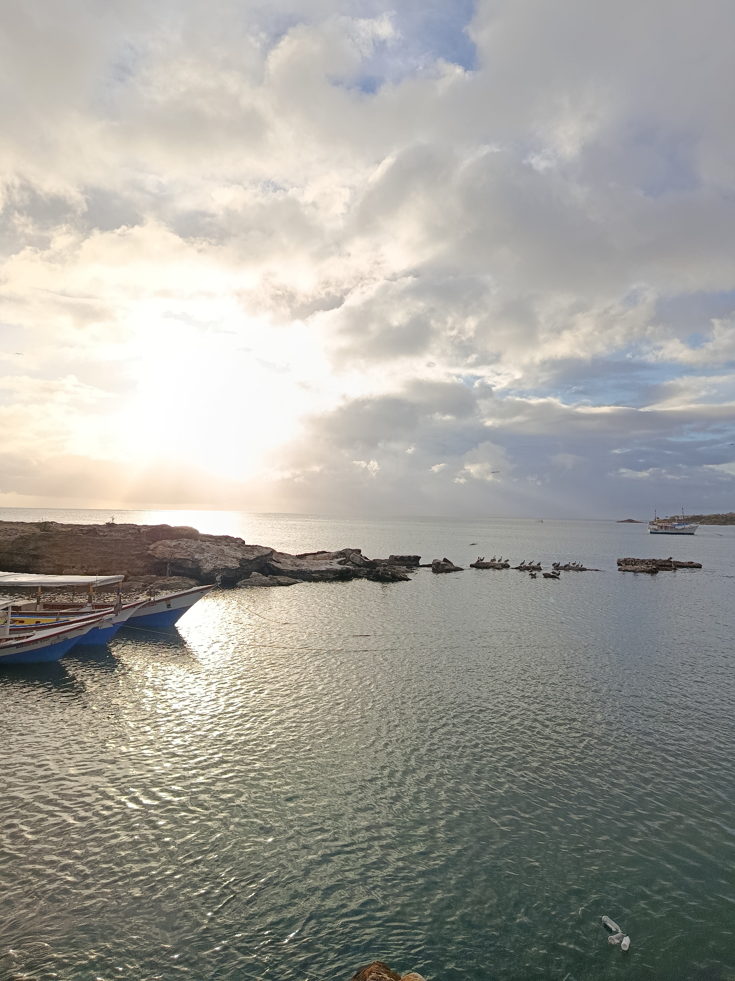 The image captures a serene coastal scene in Juan Griego, Venezuela, likely during sunrise or sunset, given the bright, low-angled sunlight illuminating the water and sky. In the foreground, several colorful boats are docked, their reflections shimmering on the gently rippling water. The boats appear to be traditional fishing vessels, with some featuring covered areas.
A rocky shoreline stretches across the middle ground, forming a natural breakwater. Scattered on the rocks are numerous birds, likely pelicans or similar seabirds, perched and observing their surroundings. Further out in the water, a larger boat is anchored, appearing to be a more substantial vessel, possibly a ferry or a tour boat. The sea is calm, with the sun creating a bright glare on the horizon, partially obscured by scattered clouds. The sky above is a blend of soft blues and whites, with the sun's rays breaking through the clouds, casting dramatic light and shadows. The overall atmosphere is peaceful and natural, showcasing a typical day in this coastal Venezuelan town.