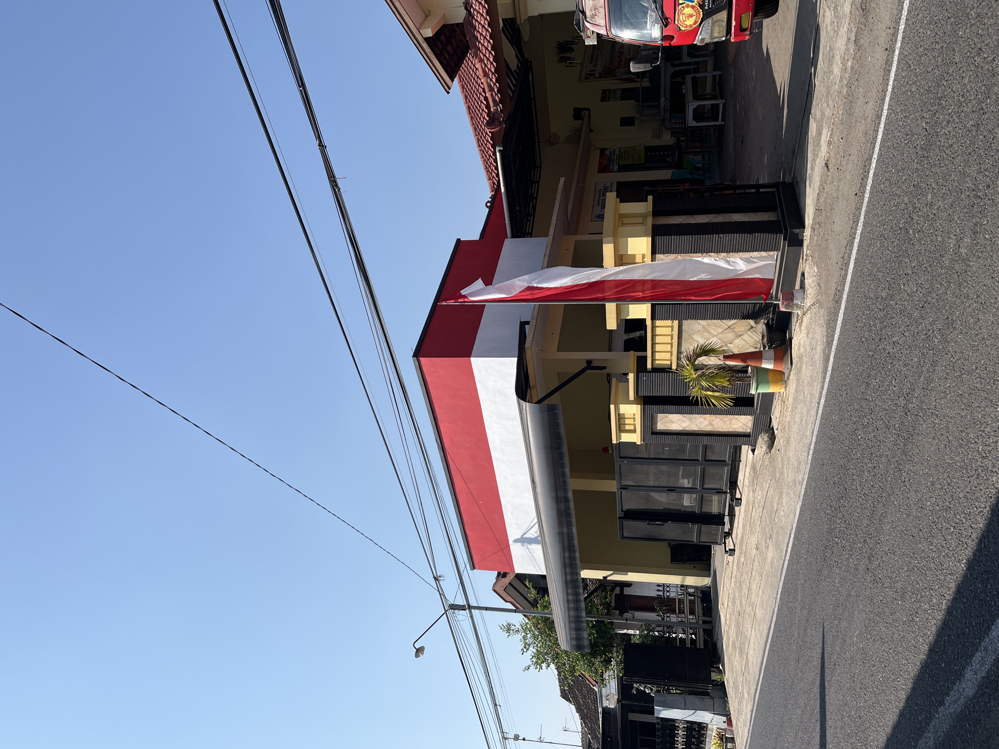 The image depicts a street scene in the city of Margorejo, Indonesia, under a clear blue sky. In the foreground, an asphalt road with a white dividing line is visible, with the shadow of a building stretching across it.
To the right of the road, a building with a red-tiled roof is prominent. The building's facade features a horizontal stripe of red at the top, followed by a white section. A black awning extends from the building. In front of this building, a tall, vertical flagpole displays the Indonesian flag, which consists of two horizontal bands: red on top and white on the bottom. The flag is partially unfurled and positioned to the left of the building entrance.
A black metal gate, appearing to be a driveway entrance, is situated in front of the building. Several large black speaker cabinets are positioned behind the gate. To the left of the gate, a lamppost with a curved arm extends over the sidewalk. Adjacent to the building and the gate, there are some potted plants, including a small palm tree, and a traffic cone.
Further to the right, a red vehicle, possibly a truck, is partially visible, suggesting it is parked or passing by. The background shows more buildings with traditional tiled roofs and some trees. The overall environment appears to be a residential or commercial street in an Indonesian town. There are no people clearly visible in the image. The scene is captured in daylight, with bright sunlight casting distinct shadows. Overhead, several dark electrical wires are strung across the sky.
