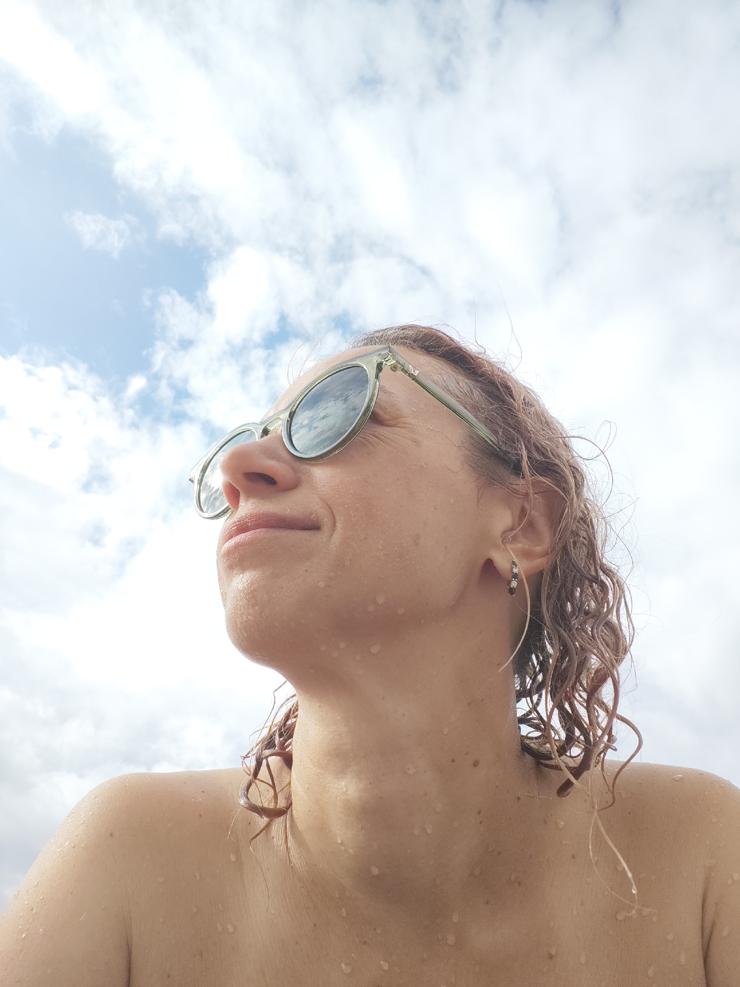 A medium shot portrait of a woman with light skin, wearing sunglasses with round, light green frames. She has curly, light brown hair that appears wet. Her skin is dotted with water droplets. The background is a sky with scattered white clouds. The photo is taken from a low angle, looking up at the woman.