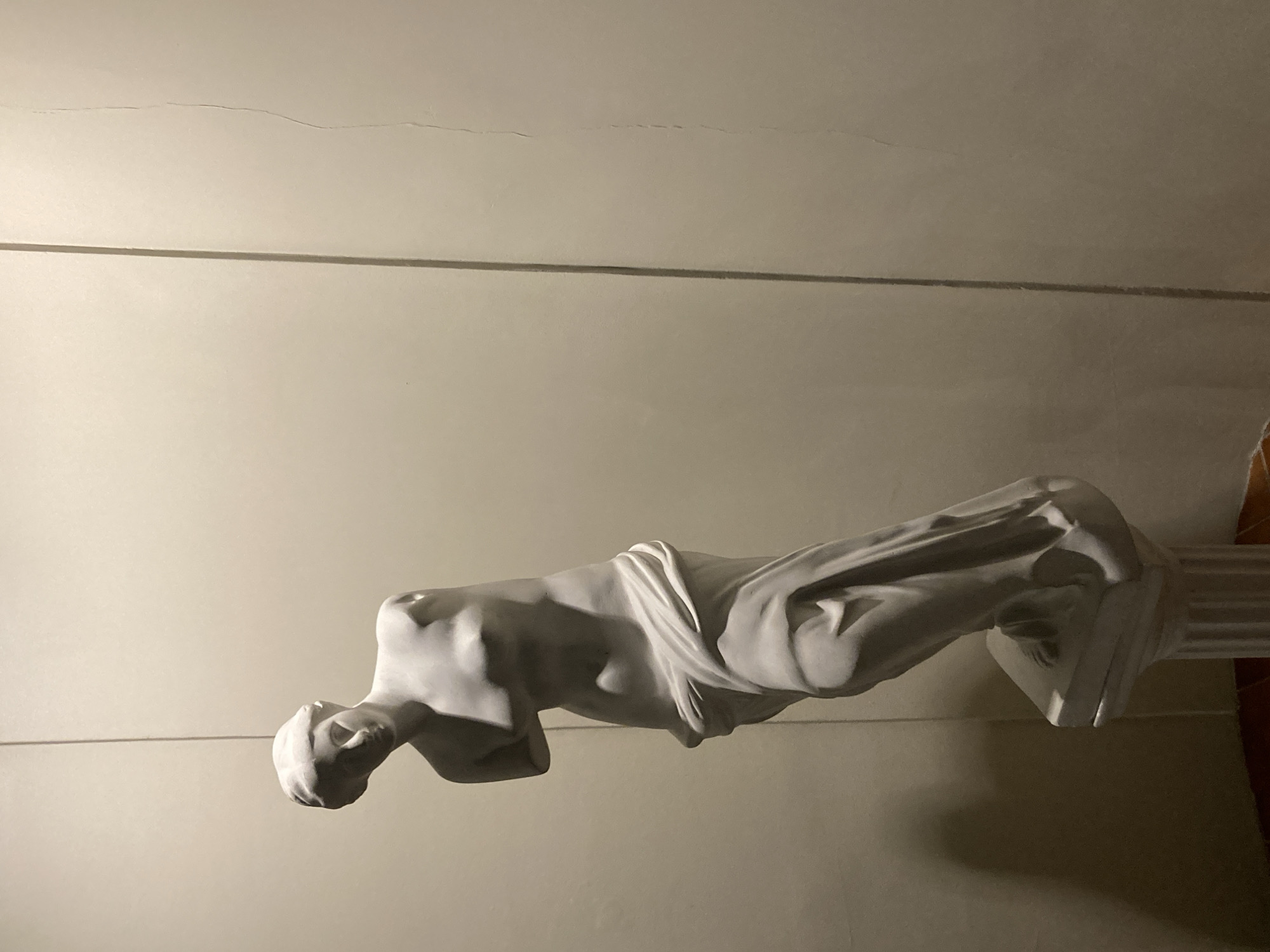 The media file presents a white, classical-style statue of a draped human figure, likely female, positioned horizontally against a plain, light-colored wall. The statue lies with its head on the left, looking upward relative to its own orientation, and its body extends to the right, resting on a partially visible, fluted pedestal or column capital.
A distinct dark horizontal line runs across the wall behind the statue, approximately in the middle of the frame. Above this line, the wall exhibits faint, wavy cracks. The overall setting appears to be indoors under dim, artificial lighting, which casts dramatic shadows and highlights the statue's smooth form and the intricate folds of its drapery. The statue's torso, arms, and head are clearly visible, showcasing the artistic detail of the piece. No specific activity is occurring, as it is a still depiction of an artwork. There is no visible text in the image.
