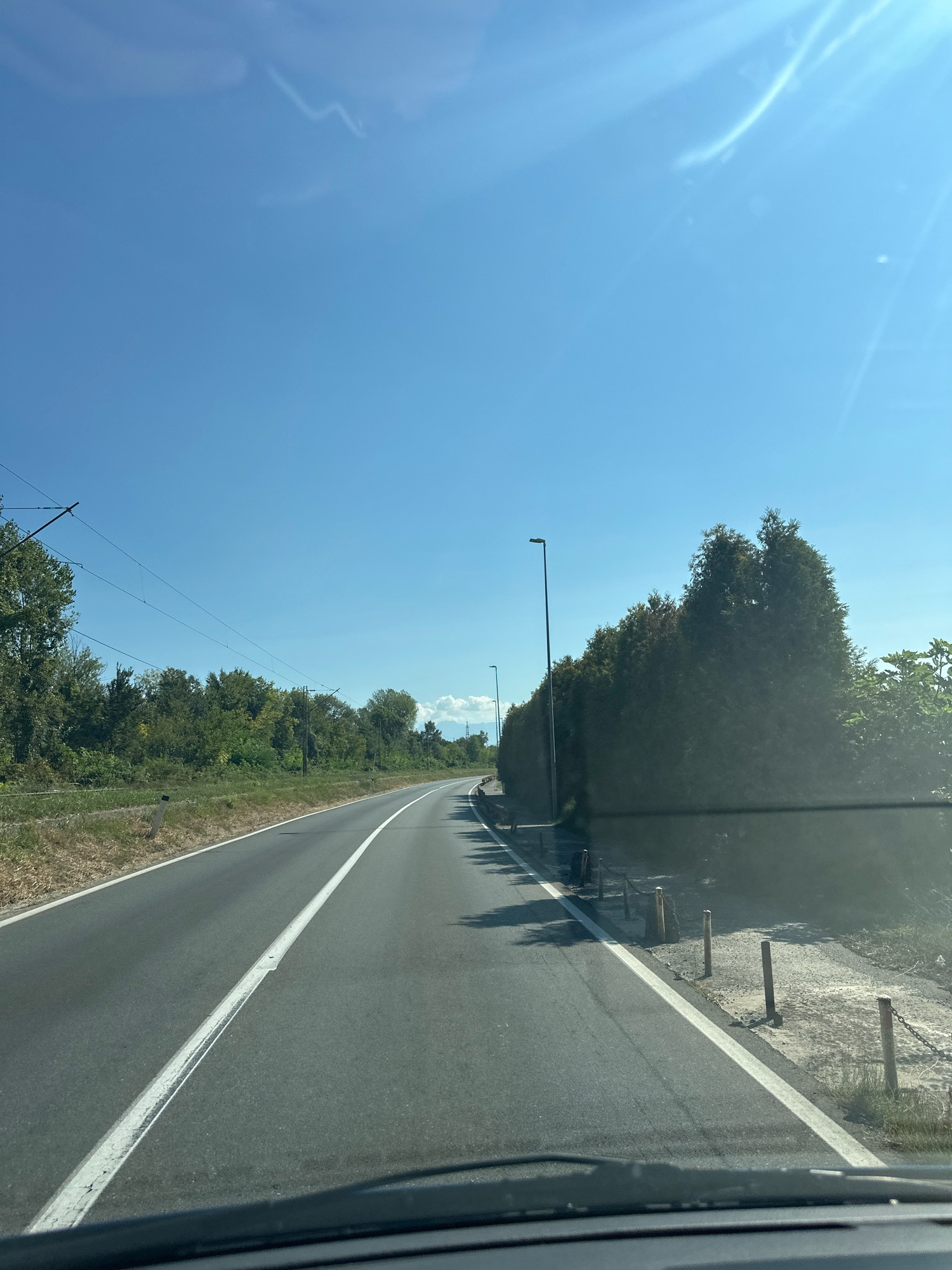 The image shows a two-lane asphalt road with a white dividing line, curving to the right as it stretches into the distance. The road is bordered on the left by a grassy verge and trees, and on the right by a row of tall evergreen trees, a sidewalk, and wooden posts connected by a chain. Overhead, power lines are visible against a clear blue sky with some wispy clouds. A street lamp stands on the right side of the road. The perspective suggests the photo was taken from a vehicle. The scene depicts a rural or suburban road on a sunny day. There are no people visible in the image.