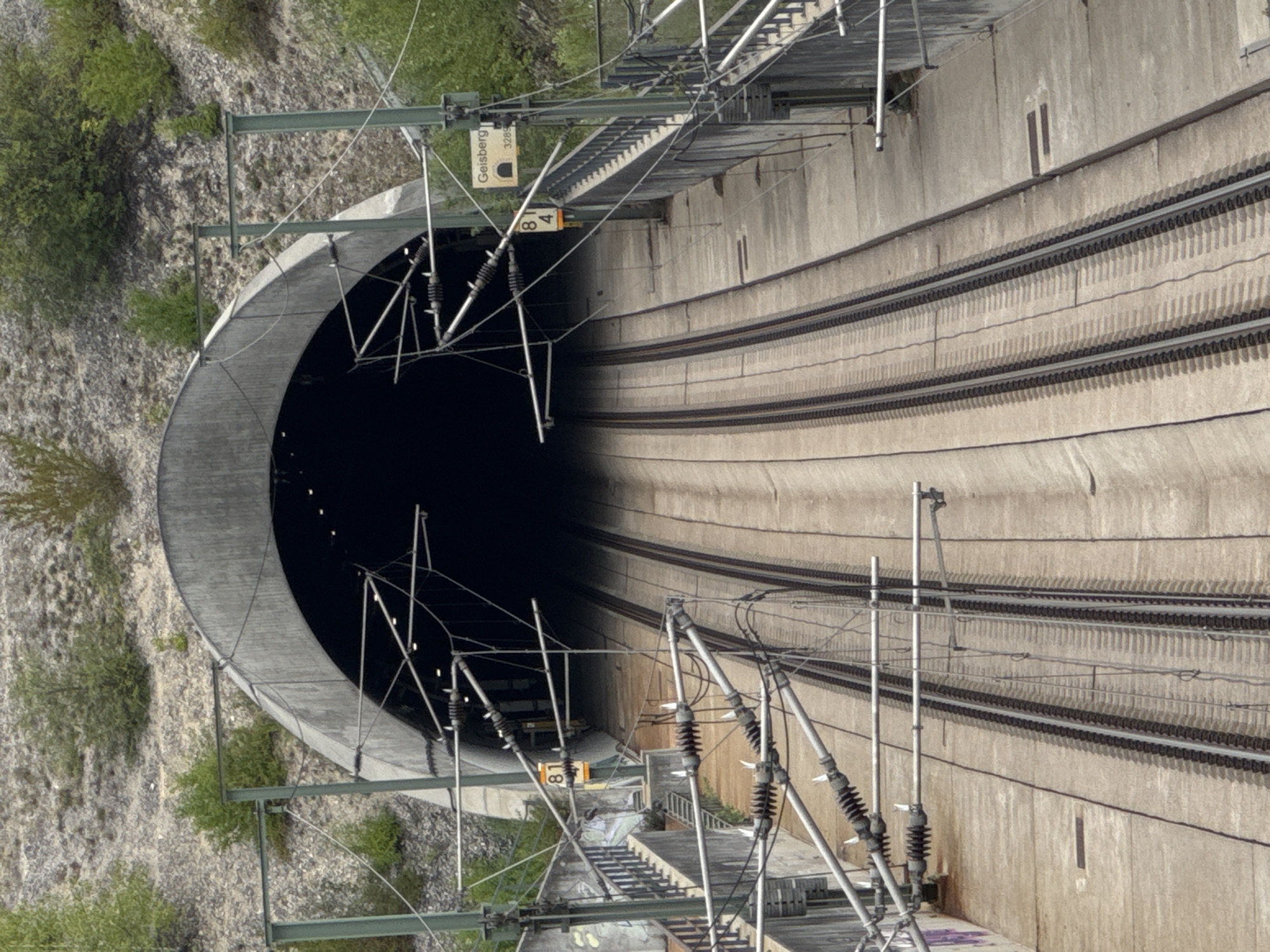 This is a high-angle, medium shot of a train tunnel entrance. The tunnel has a concrete arch and appears to be dark inside. Two sets of train tracks lead into the tunnel, made of concrete and gravel. Overhead, there are multiple power lines supported by metal poles.
On the right side of the tunnel entrance, a sign reads "Geisberg" and "3289" below it, indicating the location. Two smaller signs with numbers "8" and "4" are also visible on poles near the entrance.
The surrounding environment consists of a rocky hillside with sparse green vegetation. The weather appears to be overcast or diffused light. There are no people or trains visible in the image. The scene is likely located in Wettstetten, Germany, as indicated by the sign.