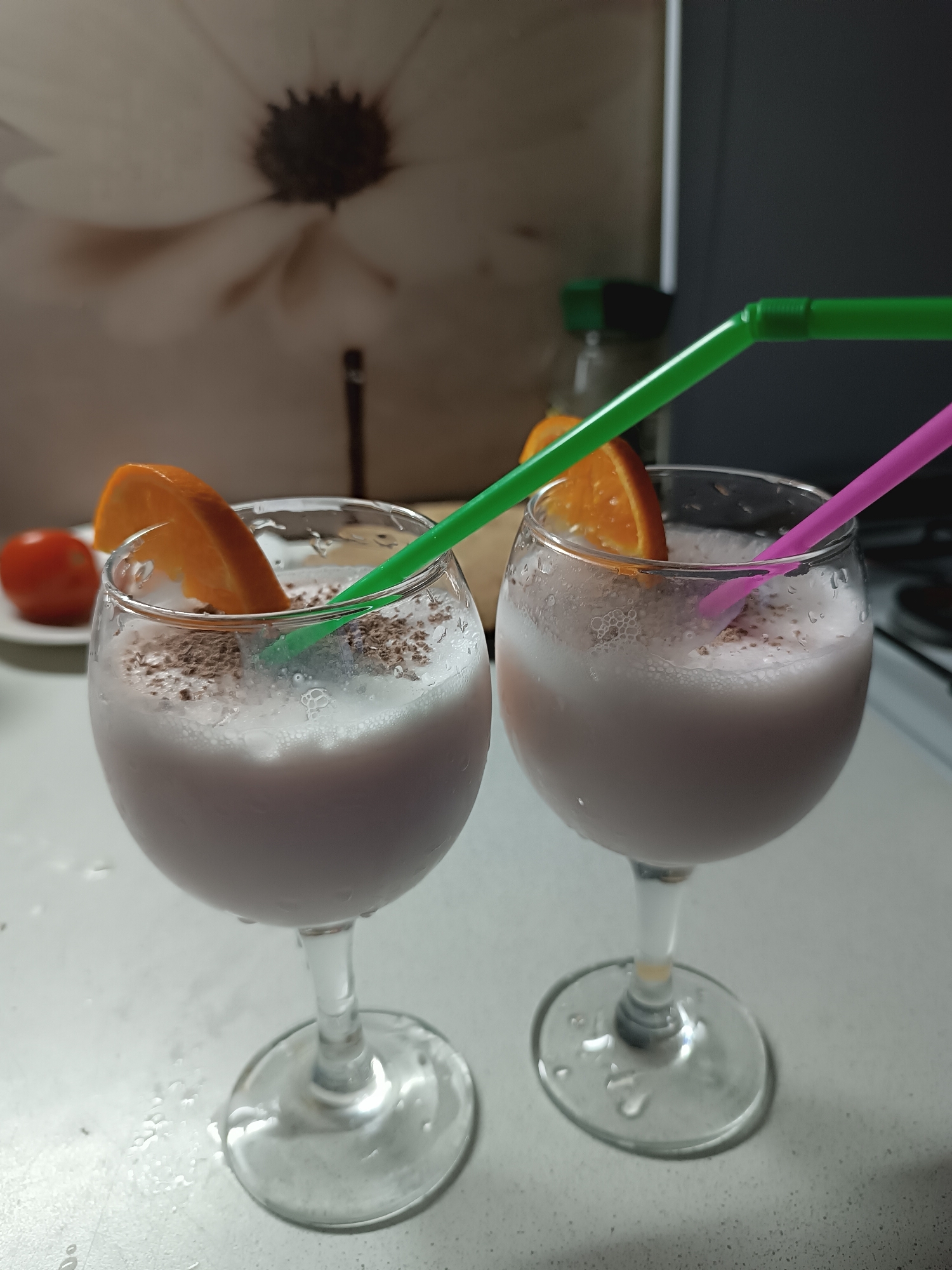 The image shows two stemmed glasses filled with a light purple beverage. Each glass is garnished with an orange slice and has a straw sticking out of the drink, one green and one pink. The beverage appears to have some kind of chocolate shavings on top. In the background, there is a tomato on a plate and a bottle with a green cap. The photo was taken indoors, possibly in a kitchen, with a countertop and wall visible in the background. A flower design is faintly visible in the upper-left corner, likely part of the wallpaper. The location is unknown.