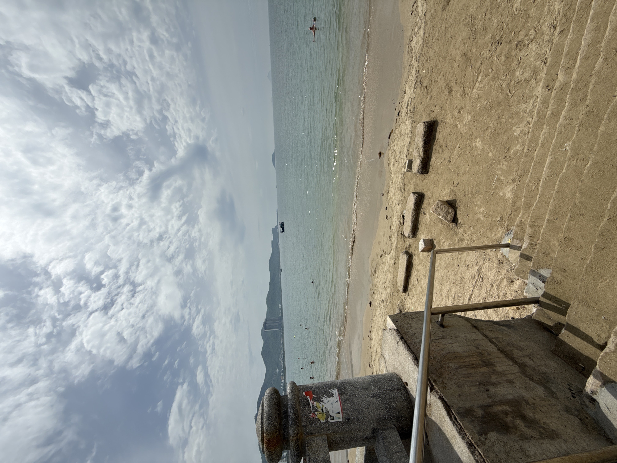 This is an eye-level, medium shot of a beach in Nha Trang, Vietnam, with the camera positioned on sandy steps leading down to the water. The sky is partly cloudy with scattered white clouds against a pale blue background. The ocean is a calm, light turquoise color, with gentle waves lapping at the sandy shore.
In the distance, a hazy coastline with green, rolling hills is visible, dotted with buildings, including a tall, modern hotel. Several small boats are scattered across the water, and a few people are swimming or wading near the shore. One person is visible floating on an inflatable raft further out in the sea.
The foreground is dominated by a stone structure with a metal railing and concrete stairs covered in sand. A concrete pillar, part of the structure, stands to the left, bearing several stickers. The beach itself is made of light brown sand and is mostly clear, with a few scattered pieces of debris and flat, rectangular stones embedded in the sand. The overall impression is a tranquil beach scene on a slightly overcast day.