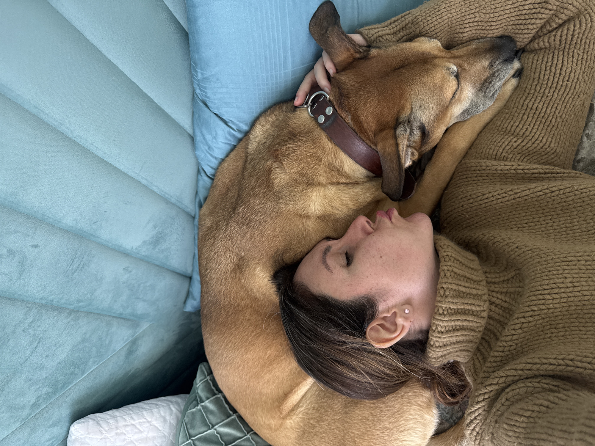 A woman is lying on a blue-green couch, closely embracing a tan-colored dog. The woman is wearing a brown sweater. The dog has a brown leather collar with metal hardware. The woman's face is visible, and she appears to be resting or sleeping. The location is unknown.
