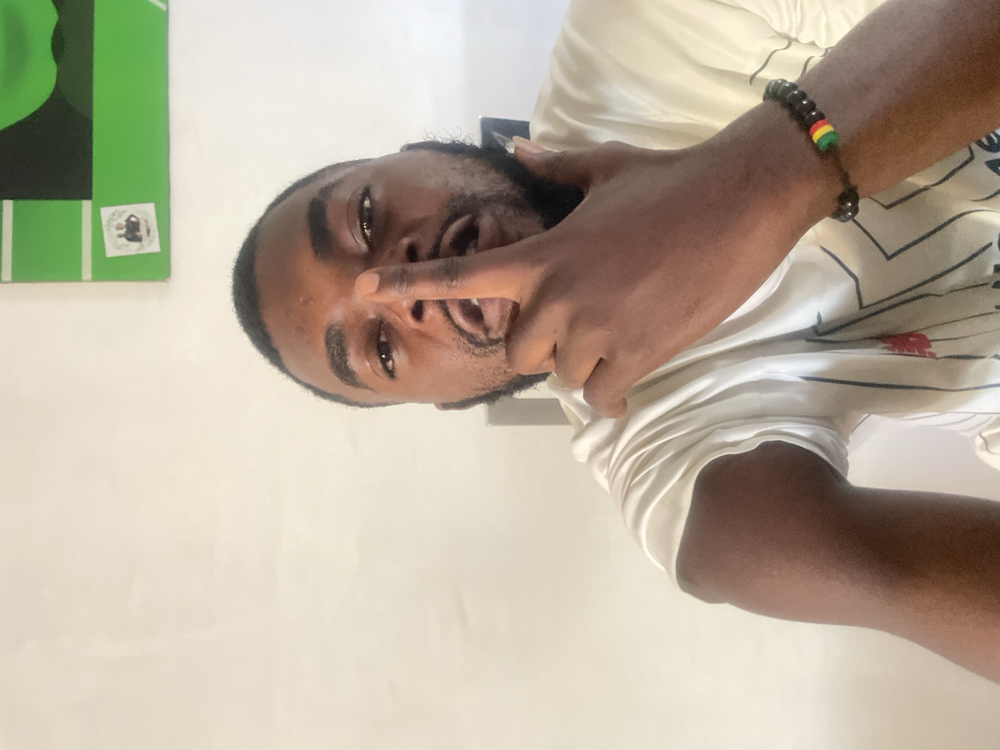 A man is visible in the image. The man has dark hair and a beard. He is wearing a light-colored shirt and a beaded bracelet with red, yellow, and green beads. The man is holding up two fingers near his mouth. A green object with white stripes and a sticker is visible in the background. It is not possible to verify the location in the city of Jos, Nigeria with the available information.