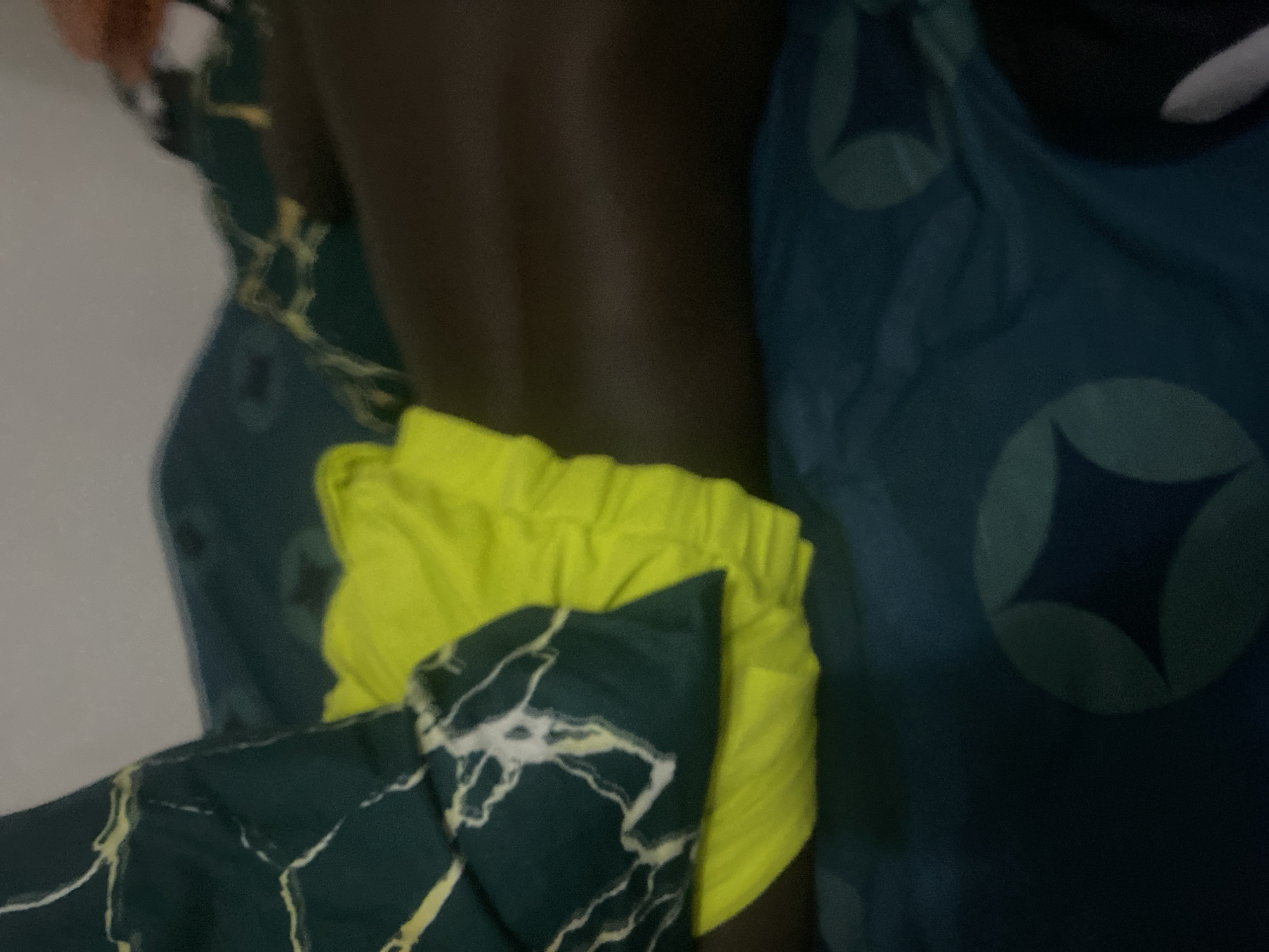 The image shows a close-up view of a person's lap with a bright yellow fabric draped over it. The fabric appears to be clothing, possibly shorts or underwear, due to its gathered waistband and texture. The person's skin is dark-toned. Behind the lap, there are bedding materials with a dark green and white marble-like pattern on one pillow and a dark blue fabric with a repeating geometric design on the other. The lighting is dim, suggesting an indoor setting, likely a bedroom. The image does not contain any information or visual cues that would allow for the identification of the location in Howayi, Nigeria.